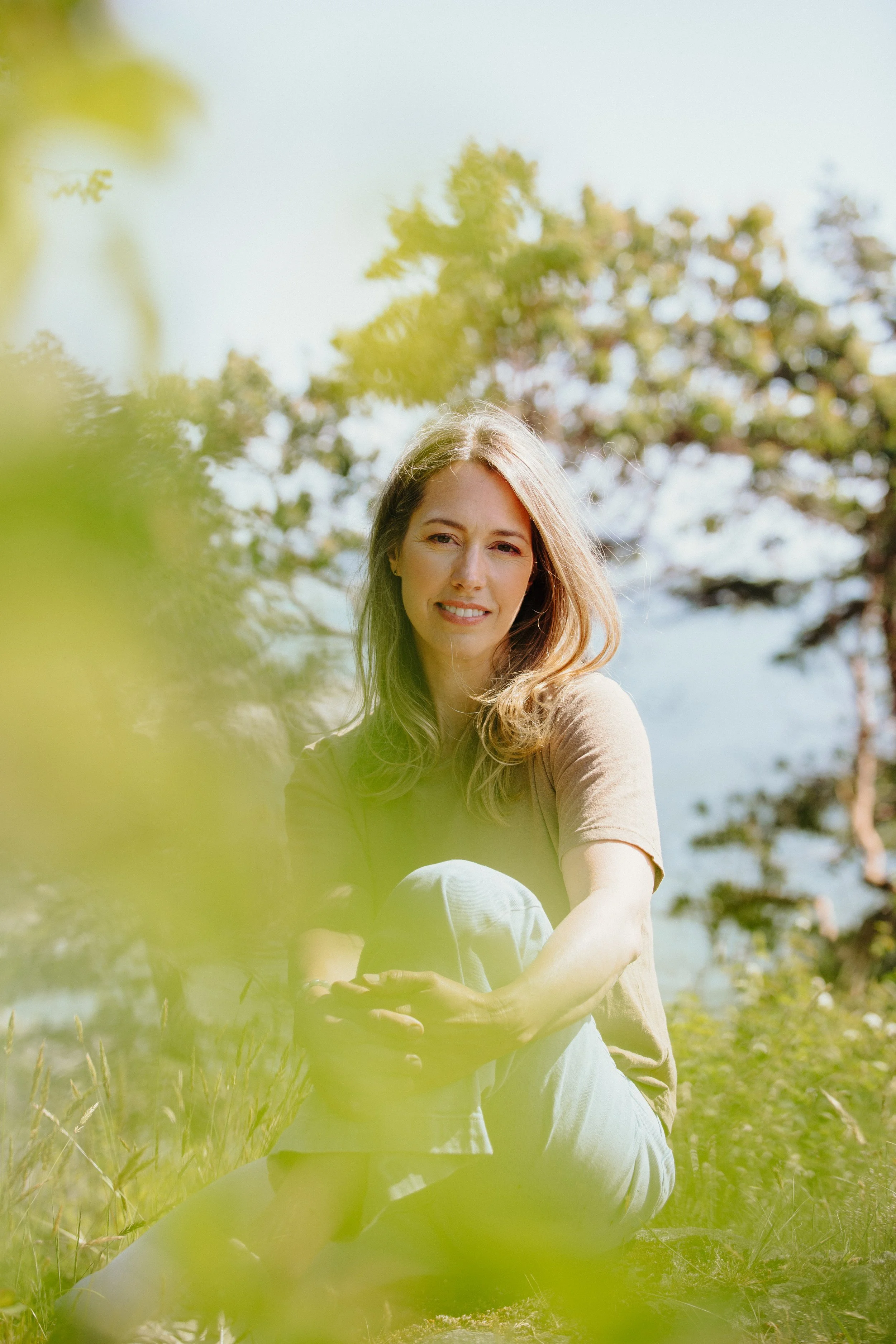 A woman with long blonde hair sitting outdoors in a grassy area, smiling, with trees and a blue sky in the background.