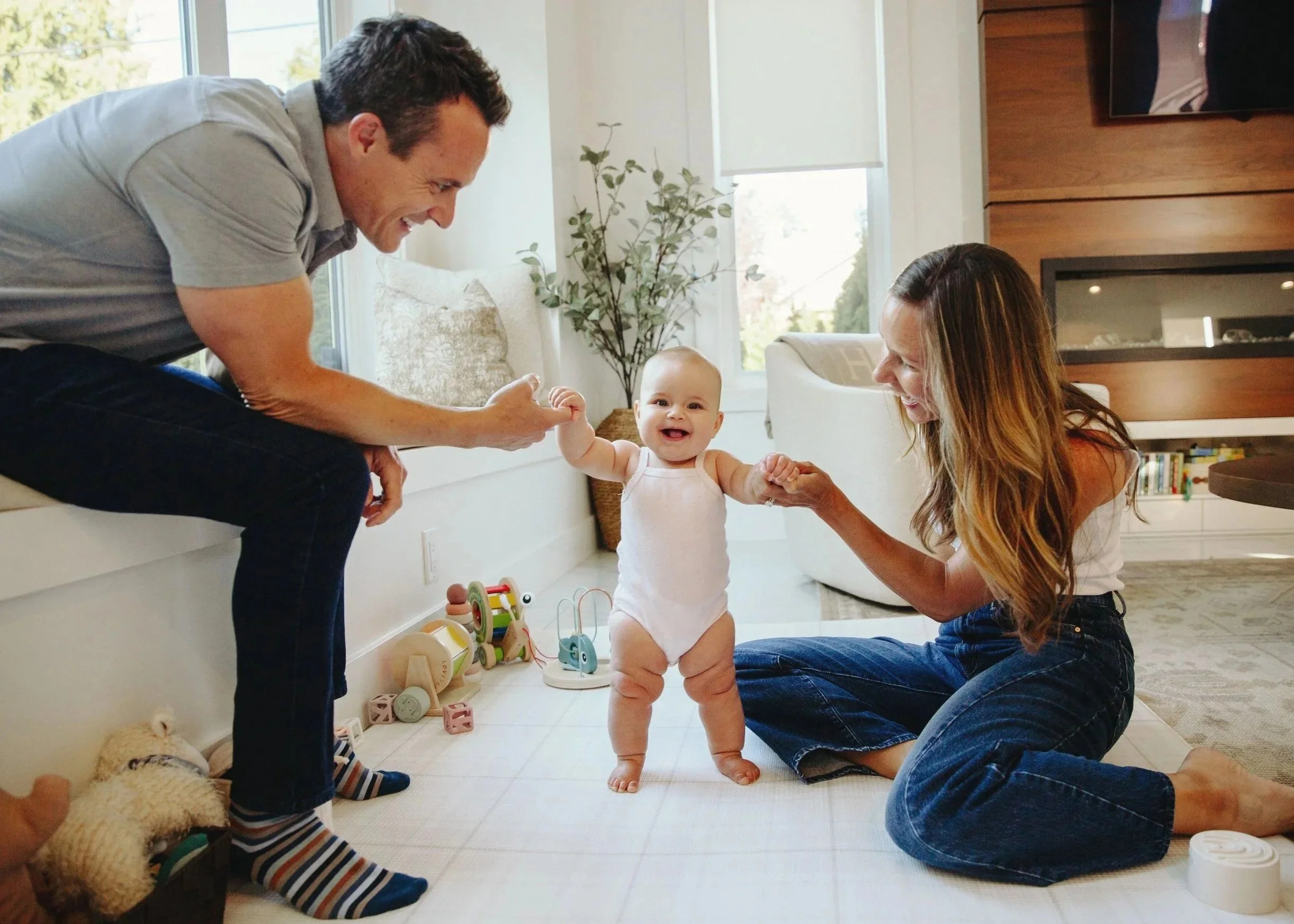 A smiling baby in a diaper standing between a man and a woman in a brightly lit living room. The man is leaning on a window sill, holding one of the baby's hands, and the woman is kneeling on the floor holding the baby's other hand. The room has toys on the floor and a picture on the wall.