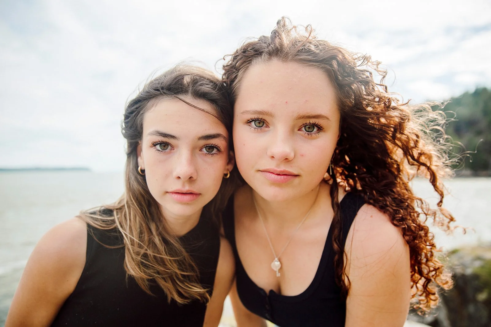 Two young women with curly hair and light skin standing close together by a body of water, with a cloudy sky and tree-covered land in the background.