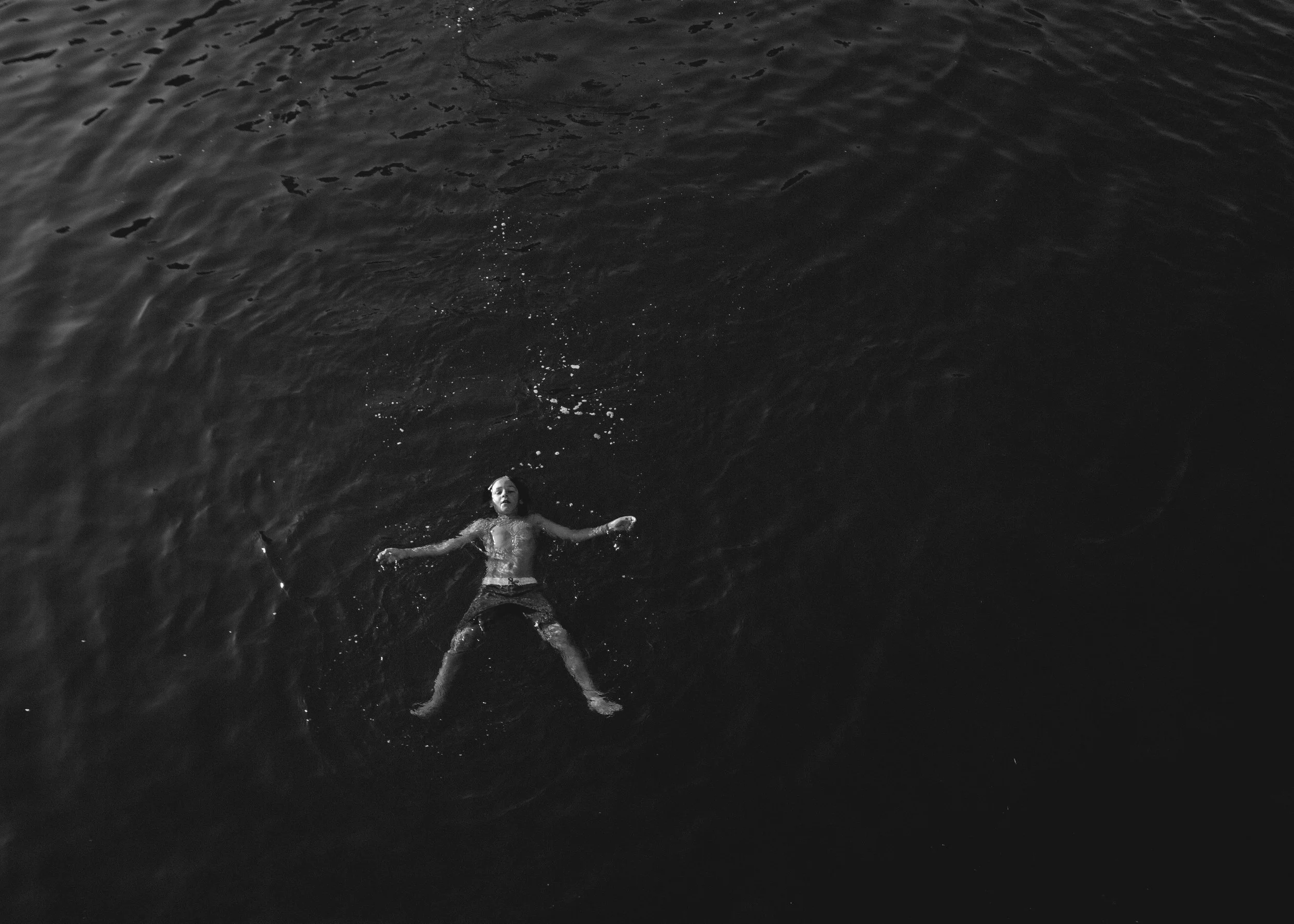 A person floating on their back in dark water during daytime, wearing swim trunks, with arms and legs spread out.