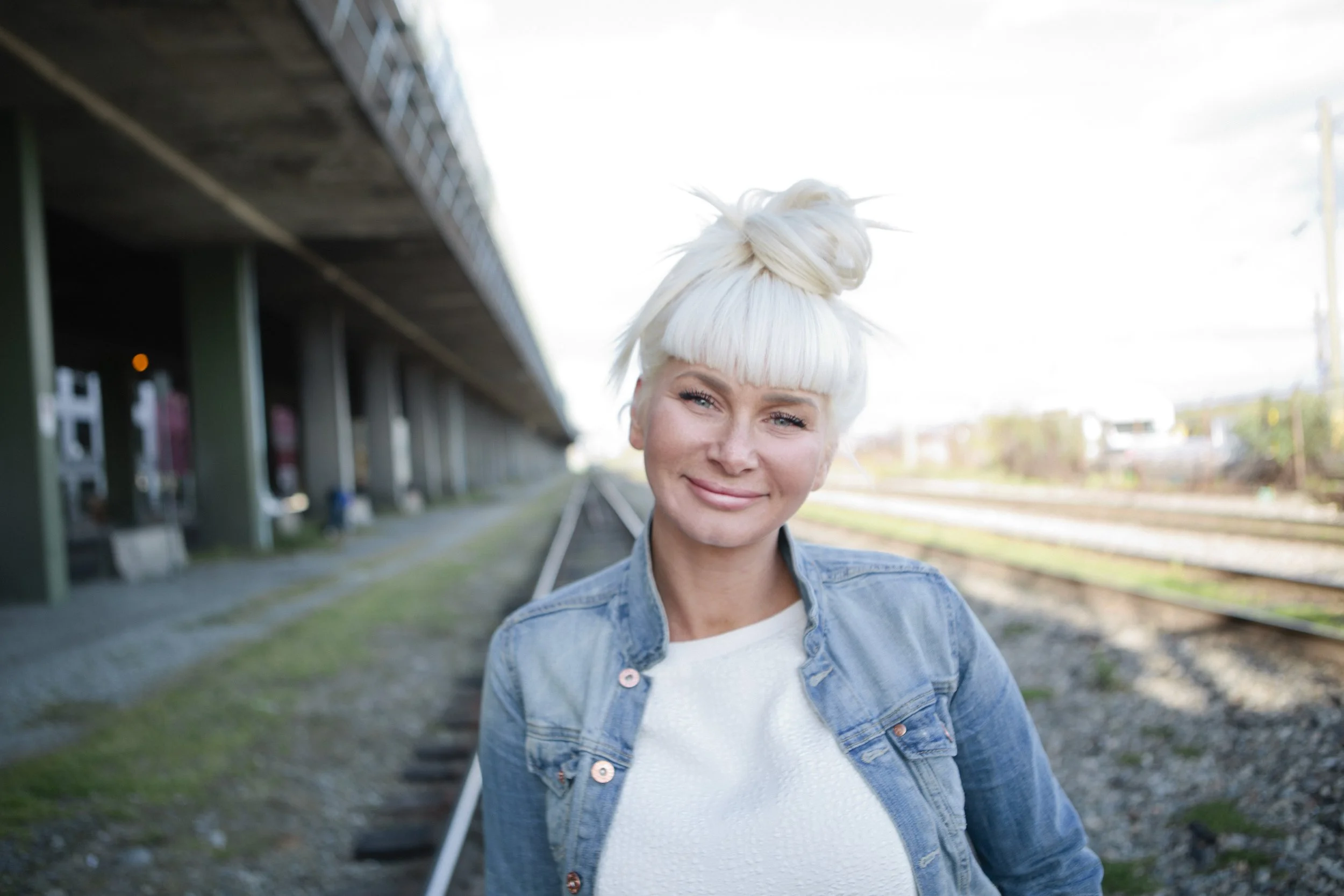 A smiling woman with platinum blonde hair tied in a top knot, wearing a denim jacket over a white shirt, standing at a train station platform under an overpass with train tracks and a cityscape in the background.