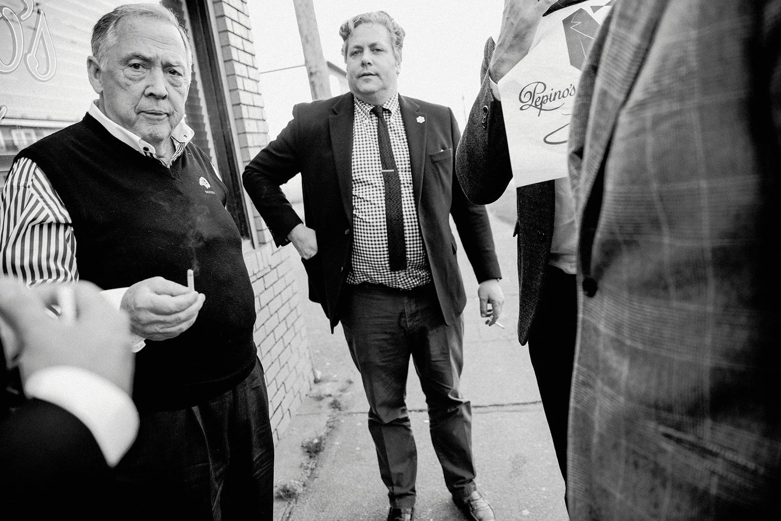 A group of men standing on a sidewalk near a brick building, engaged in conversation. One man, holding a cigarette, is in the foreground, while two others, wearing suits, are visible in the background.