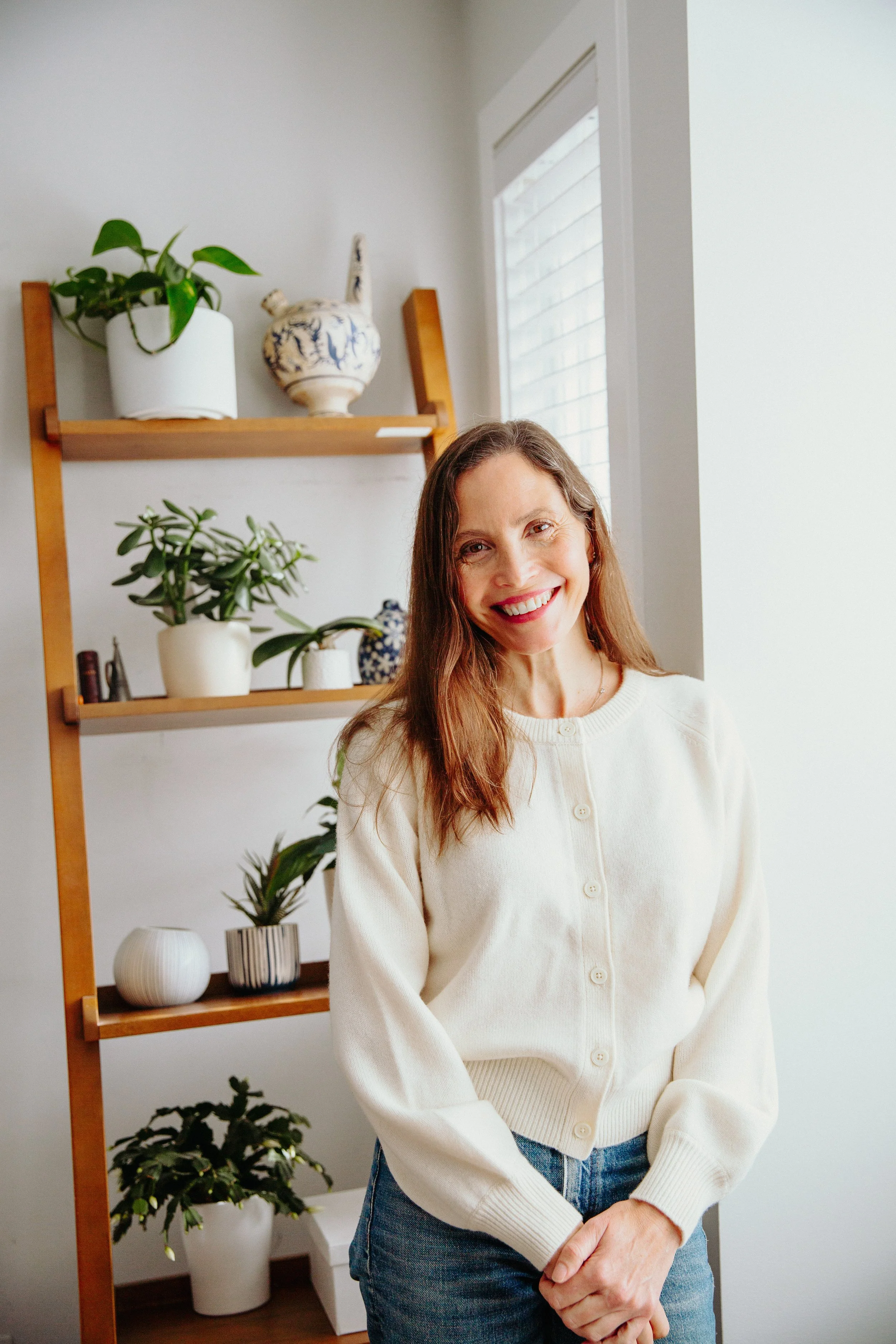 A woman with long brown hair smiling and standing indoors in front of a wooden shelf with potted plants and decorative vases, next to a window with white blinds, wearing a cream-colored cardigan and blue jeans.