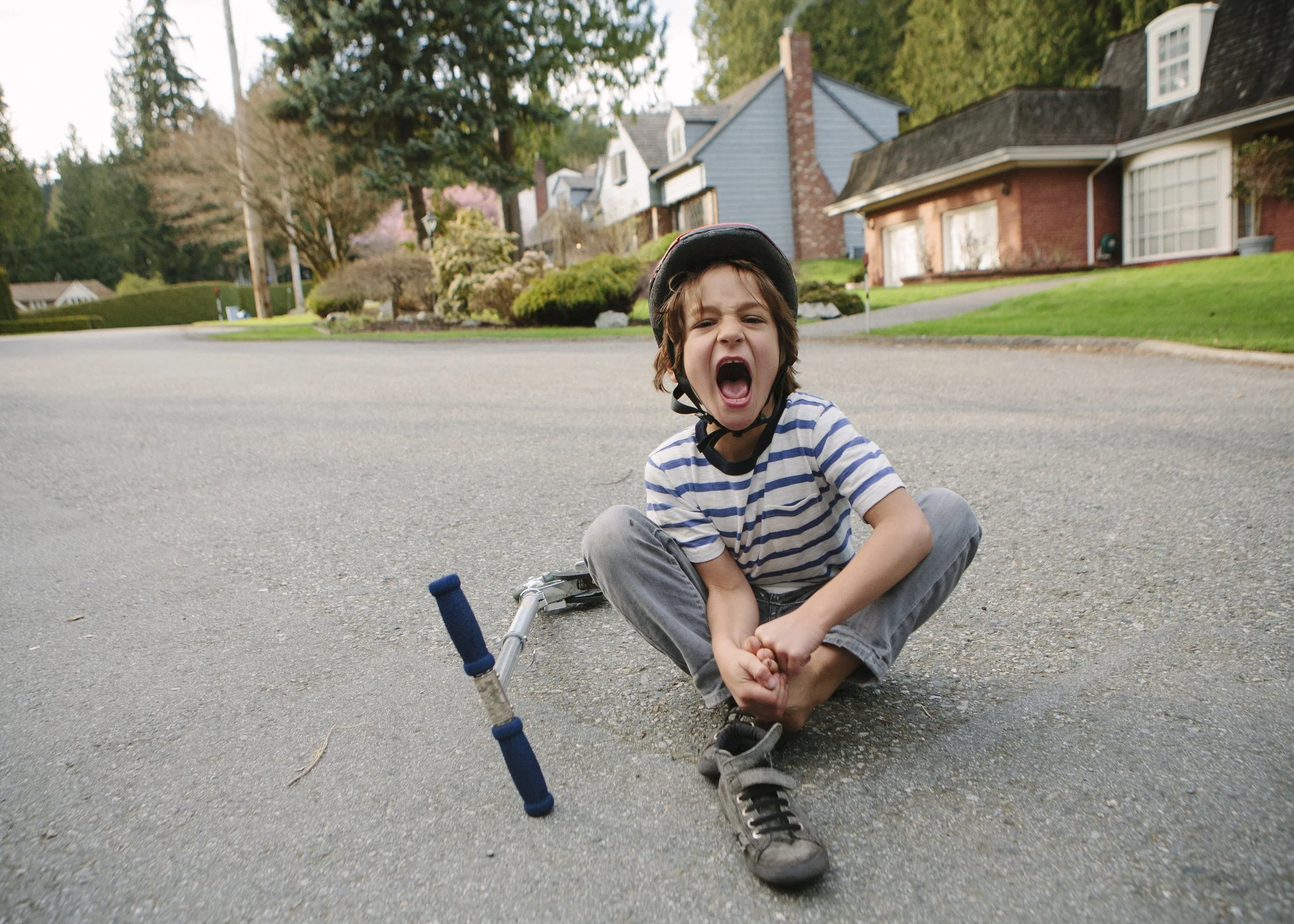 Child sitting on the pavement with a bicycle, wearing a helmet, yelling with mouth open, in a suburban neighborhood.