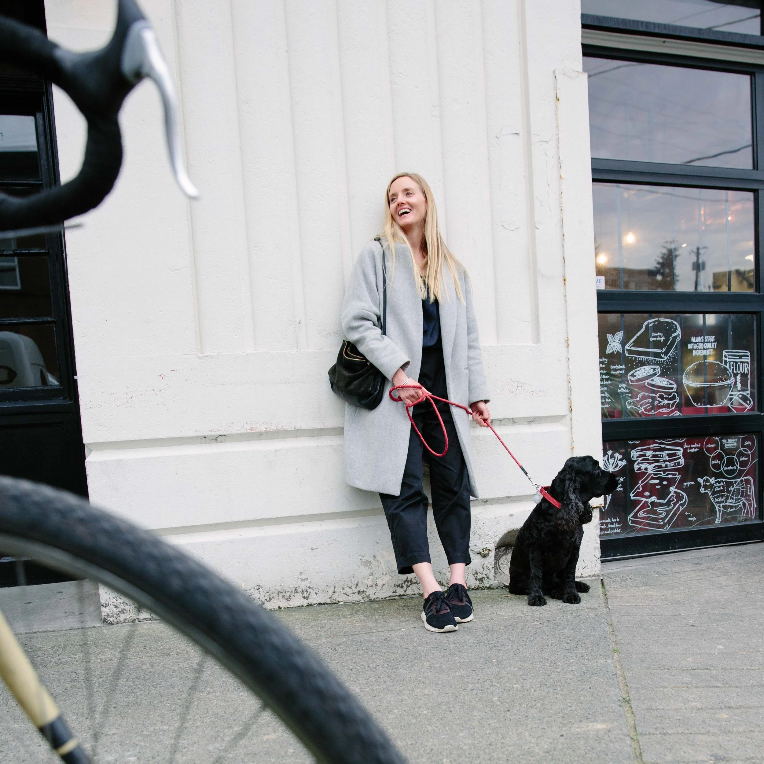 A woman with blonde hair wearing a gray coat and carrying a black bag is standing outside next to a white wall, holding a red leash attached to a black dog, with a black-framed window and bakery menu in the background.