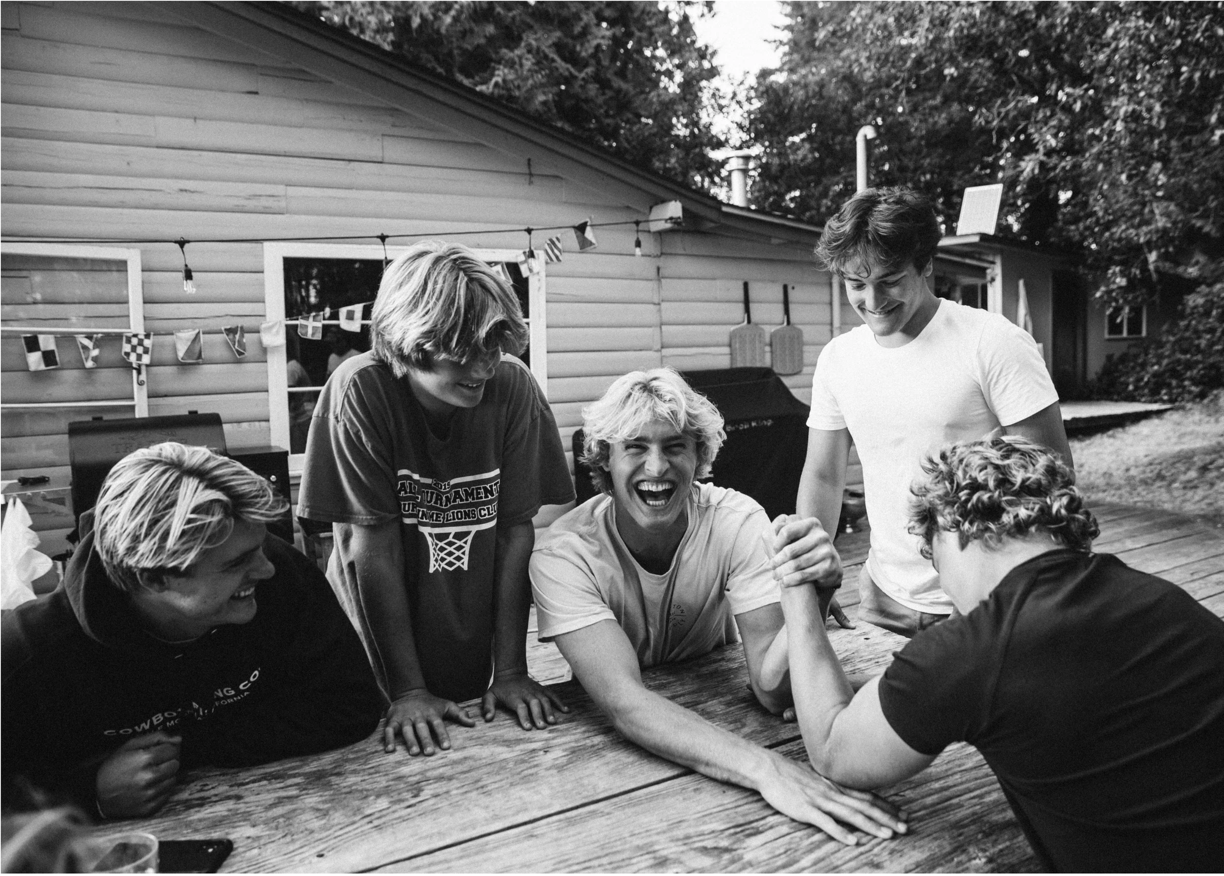 A group of young people gather around a table outdoors, engaging in arm wrestling, smiling and laughing, with a house and trees in the background.