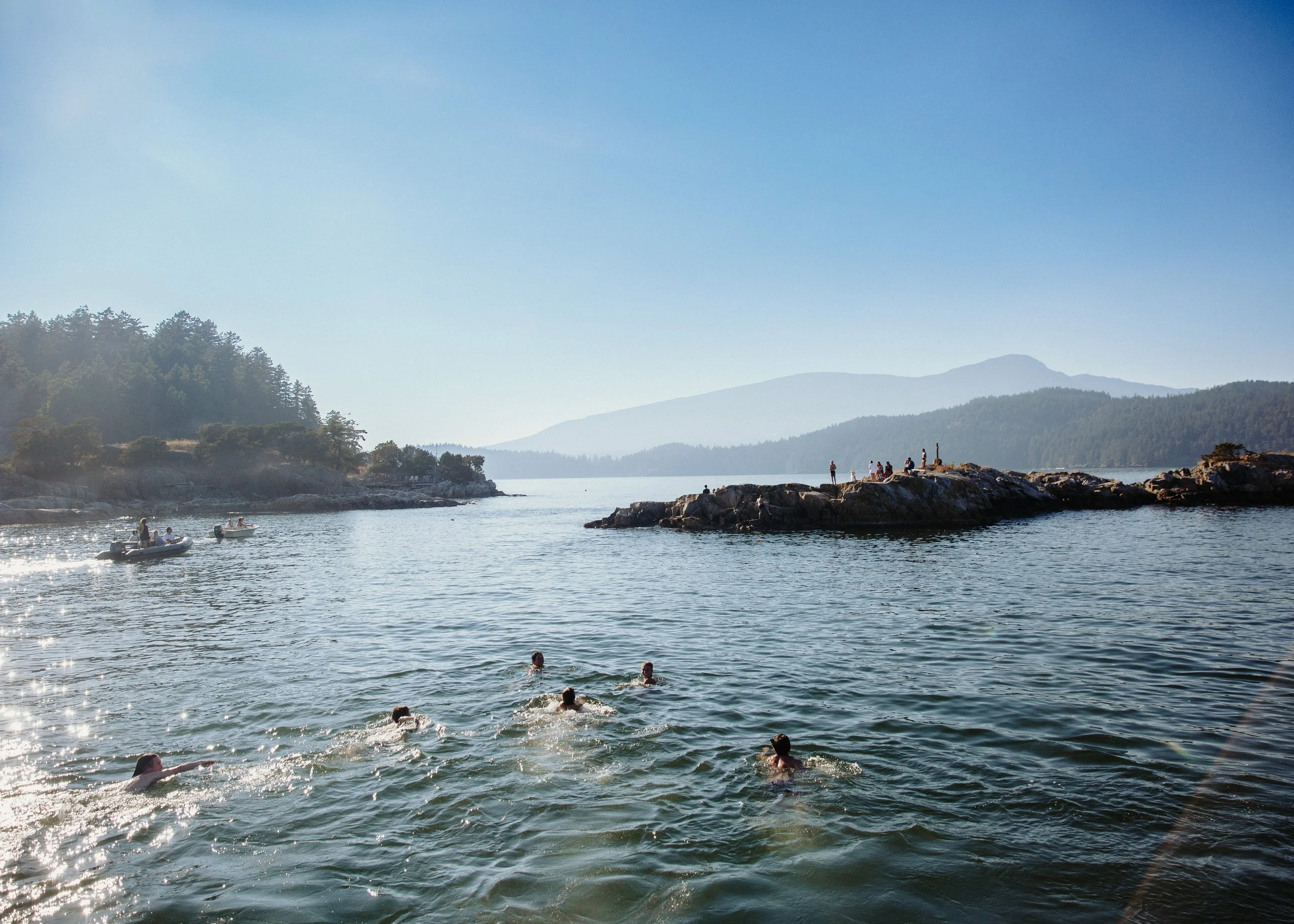 People swimming and boating in a lake with forested hills and mountains in the background on a sunny day.
