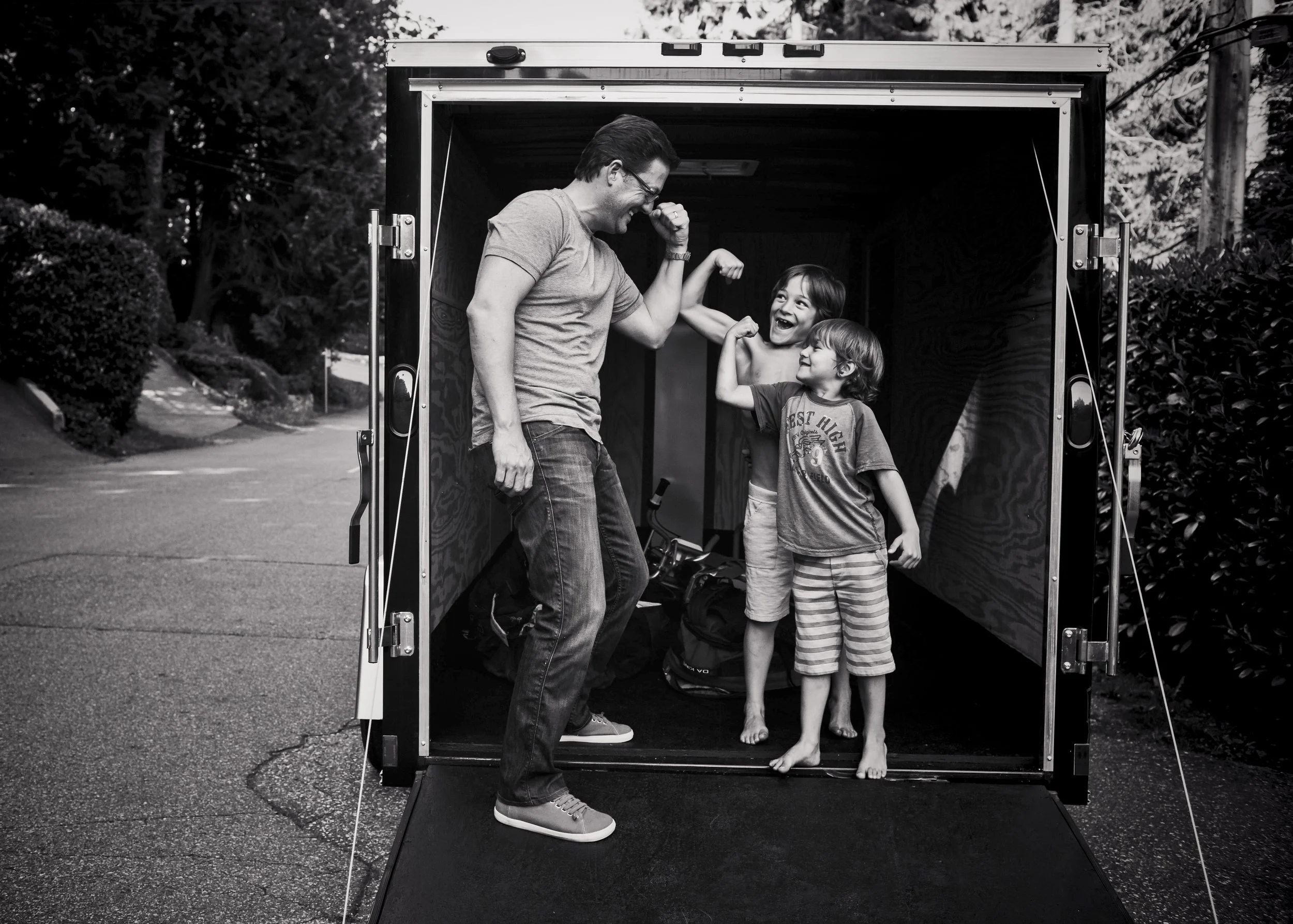 A man and two children inside a trailer, playing and pretending to flex their muscles, outdoors on a paved road with trees and bushes nearby.