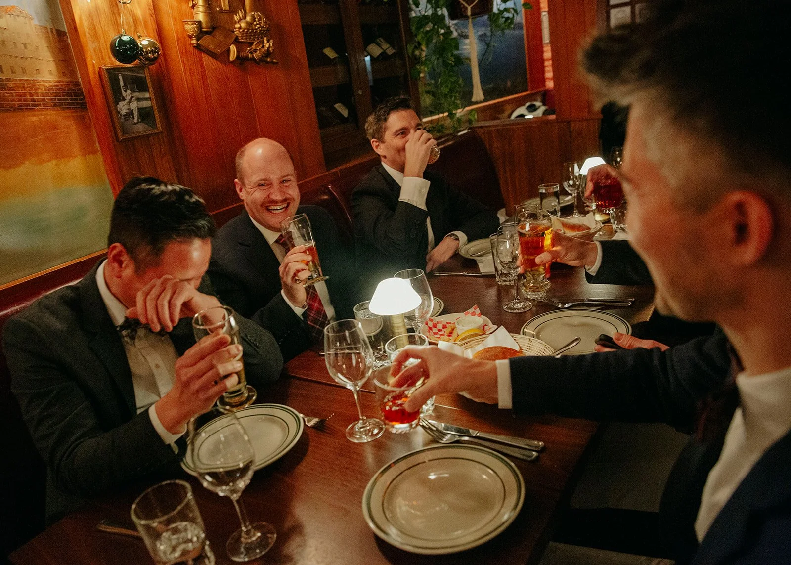 A group of men in formal attire sitting around a dinner table in a restaurant, smiling and raising glasses for a toast.