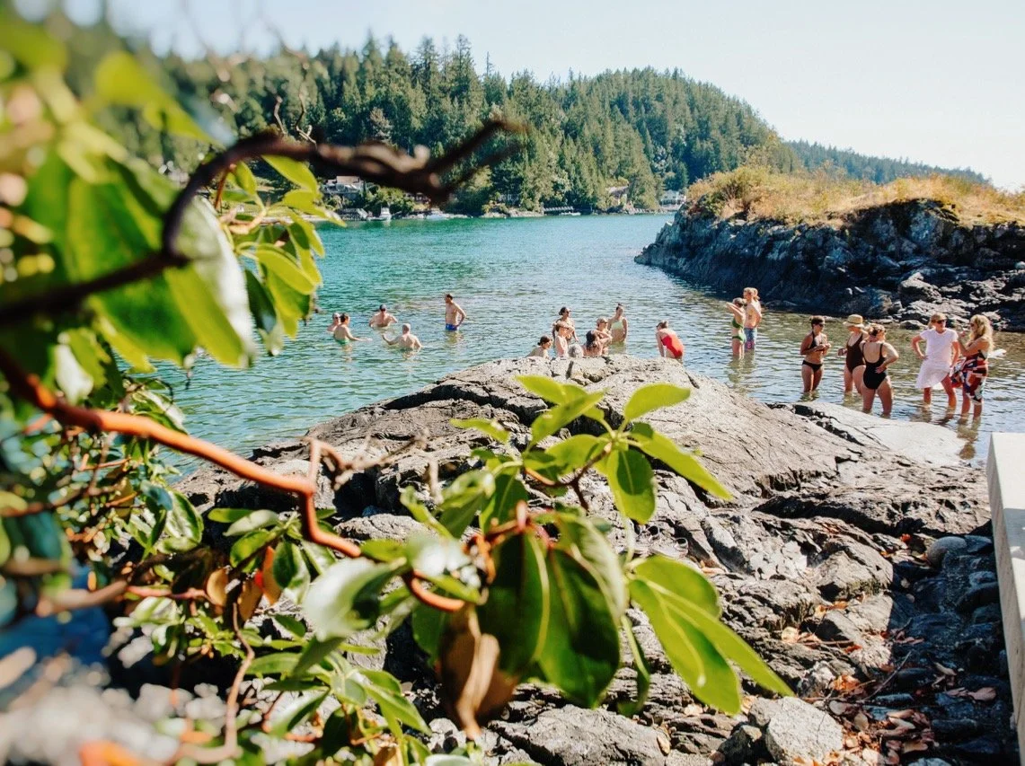 Group of people swimming and relaxing at a rocky beach with green trees in the background.