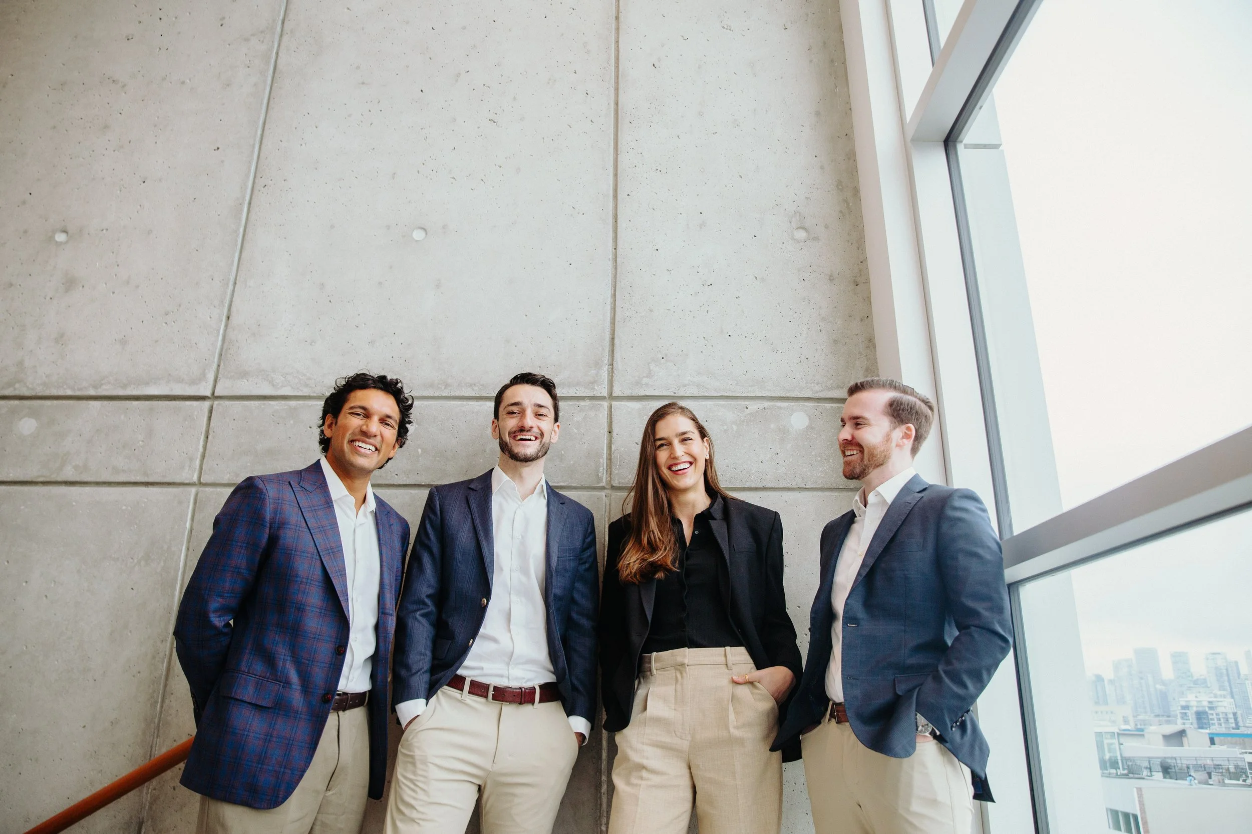 Four smiling businesspeople in suits standing inside a modern building with large windows and city views.