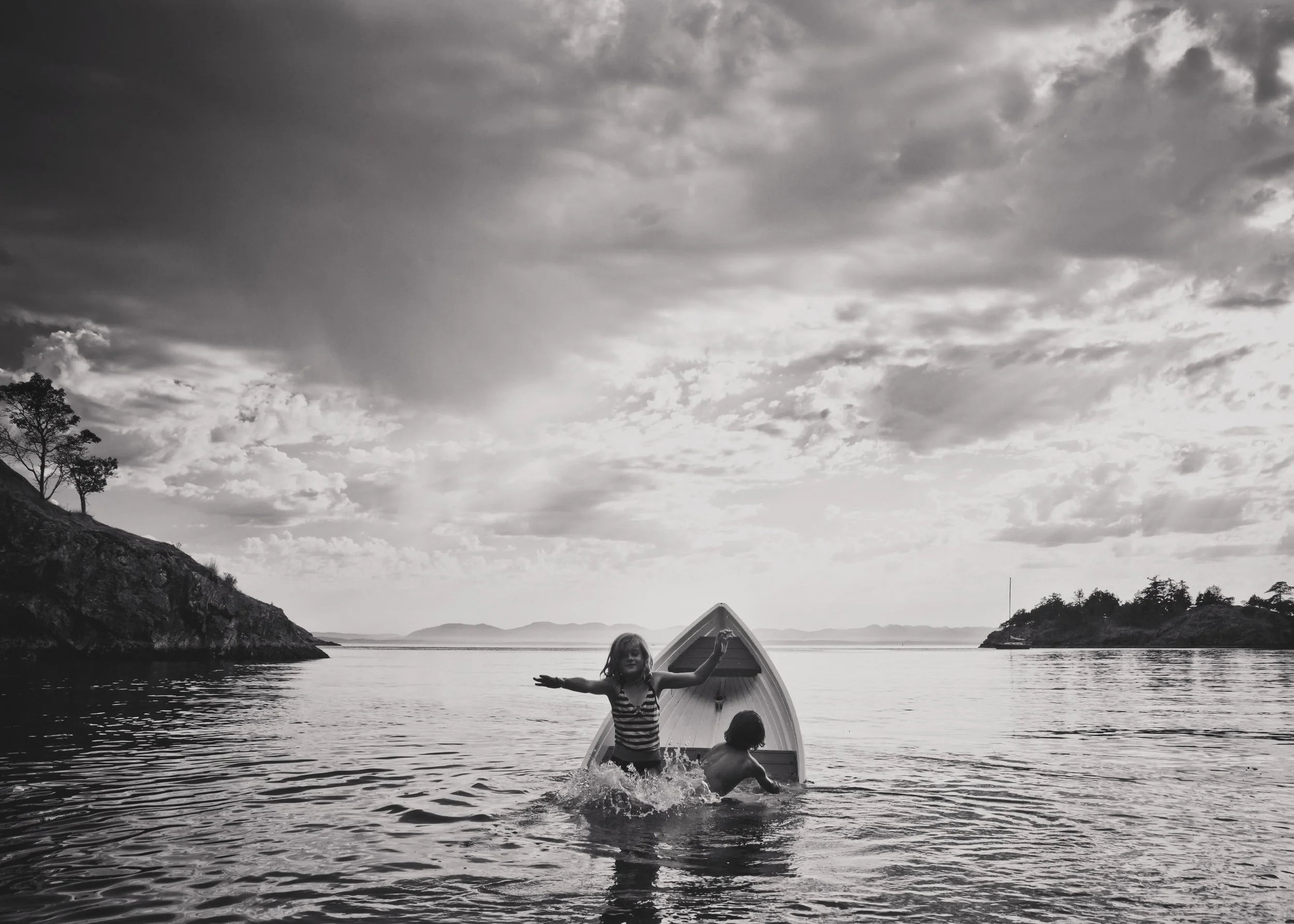 Children playing on a boat in a calm lake surrounded by mountains under a cloudy sky in black and white.