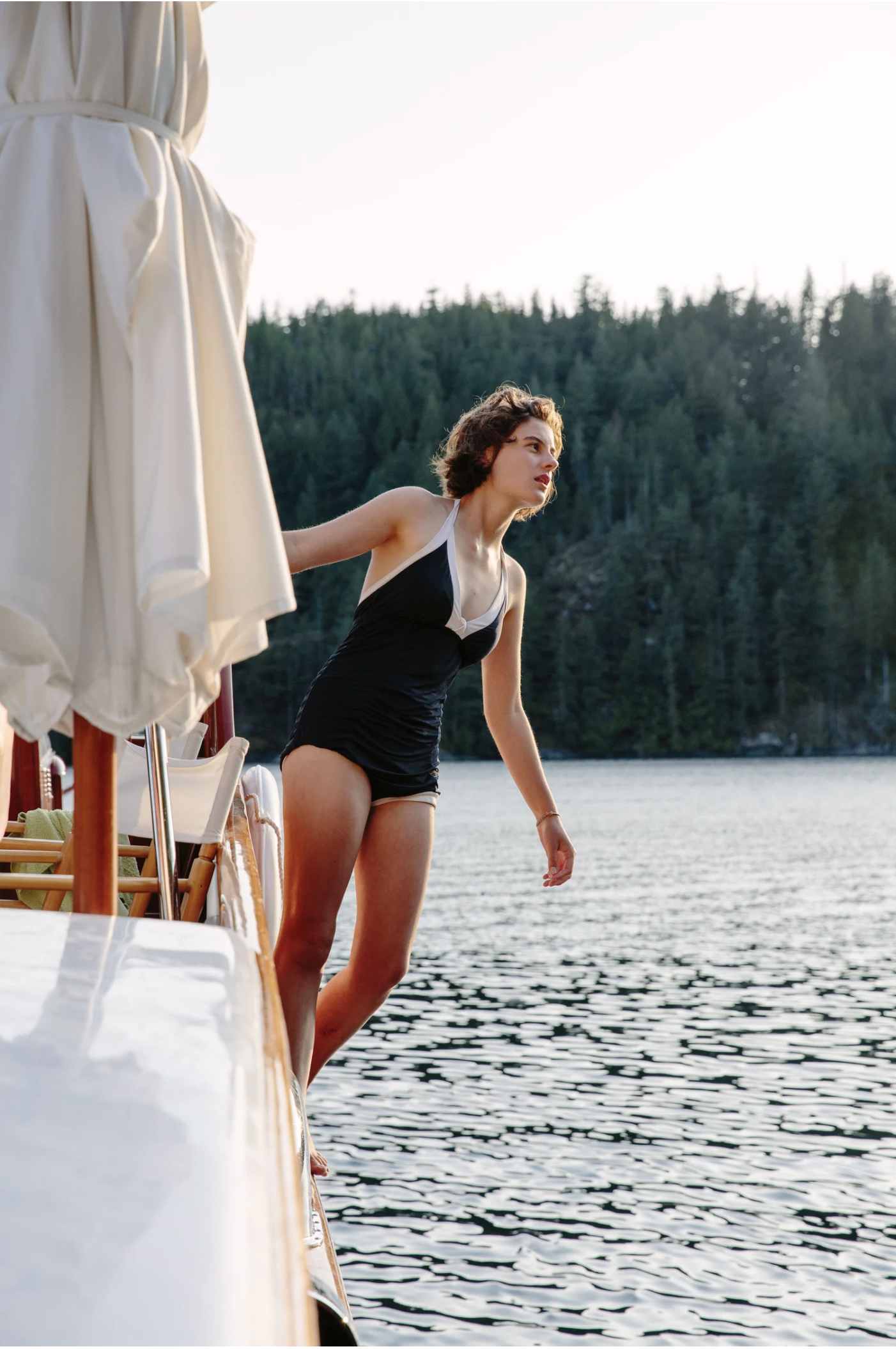 A woman in a black swimsuit stands on a boat by a lake, with a forested mountain in the background during sunset.