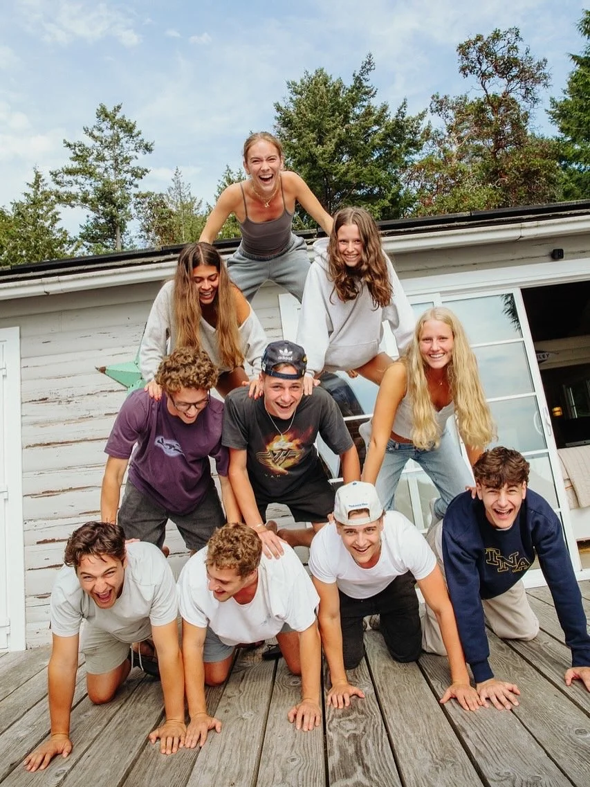 A group of nine young people forming a human pyramid outside a white wooden building with trees in the background, smiling and having fun.
