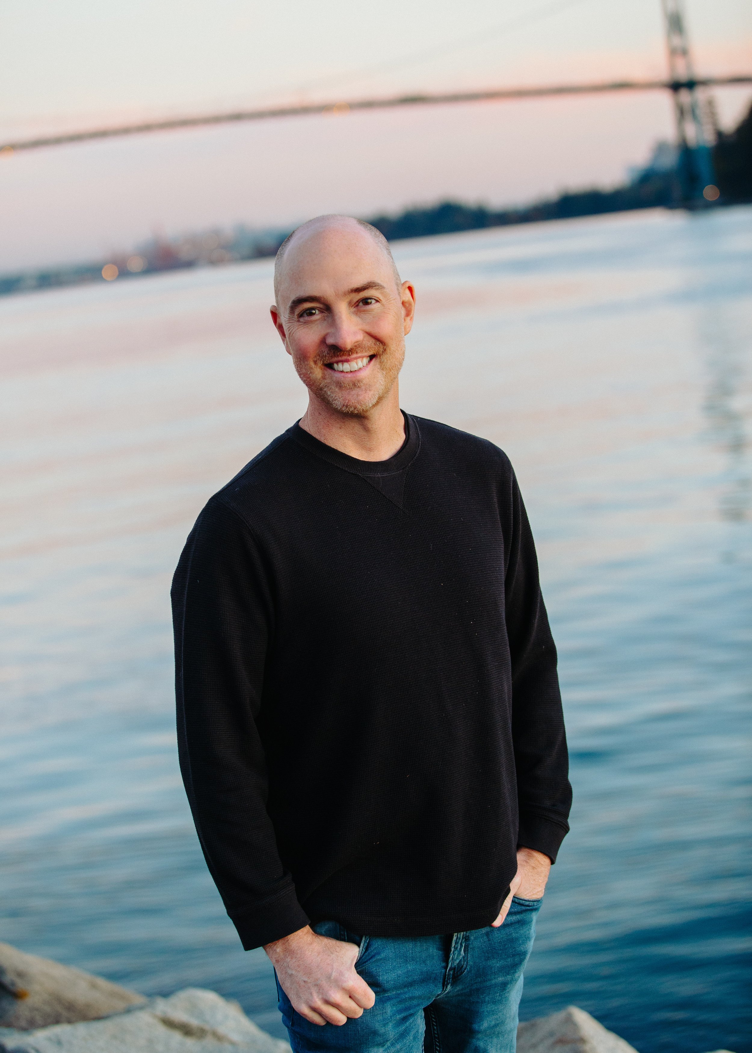 A smiling man with a shaved head and beard standing by the water during sunset, wearing a black sweater and blue jeans.