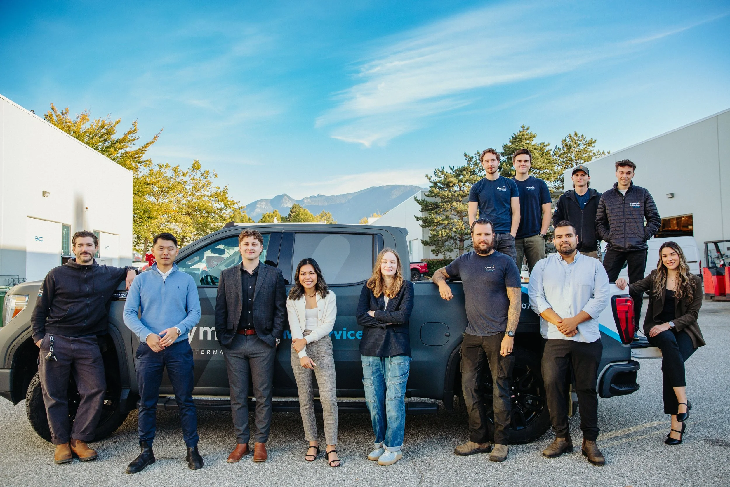 A diverse group of twelve people, including men and women, standing outdoors in front of a black service vehicle with mountains and trees in the background. They are smiling and dressed in casual and business casual attire.