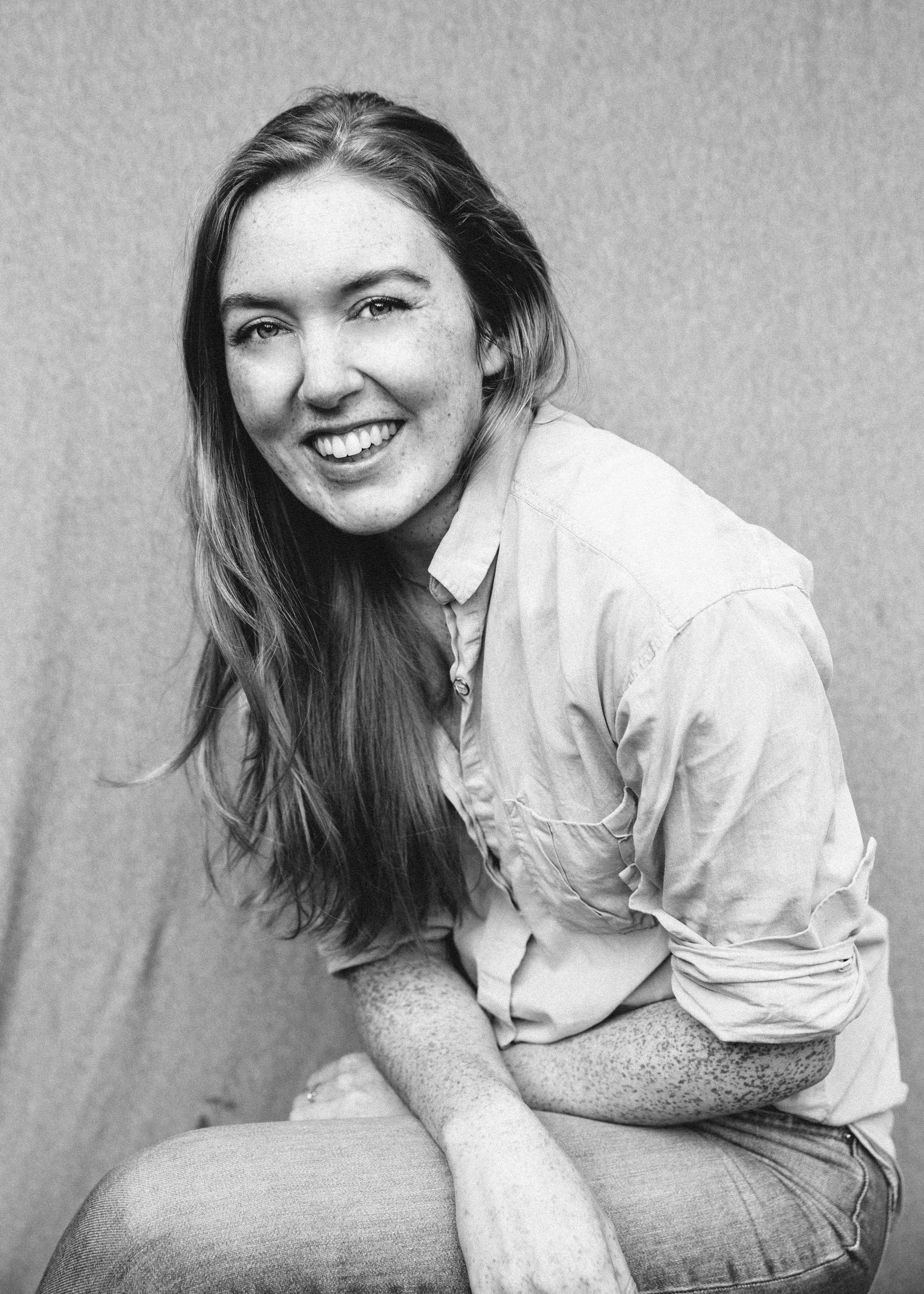 Black and white photo of a woman with long hair, smiling, and looking at the camera. She is wearing a light-colored button-up shirt and leaning on her knee, with a textured background.