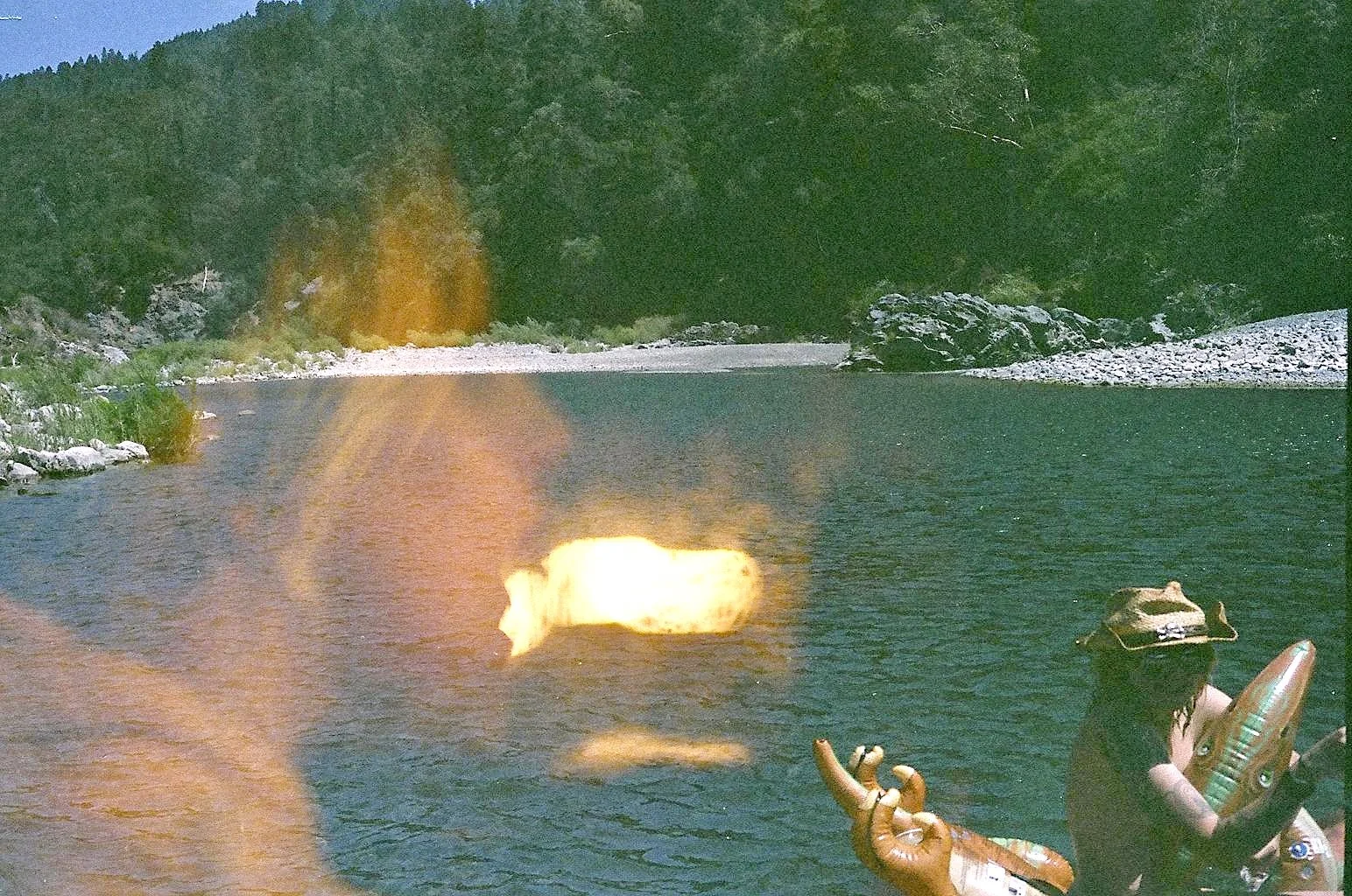 A person wearing a hat and sunglasses sitting in a boat on a lake, holding a floatation device, with a dog sitting at the front of the boat, and trees and rocky shoreline in the background.