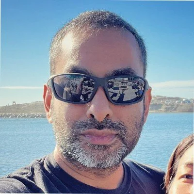 Man with short hair, beard, and sunglasses taking a selfie outdoors near water with a boat and distant shoreline visible in the background.