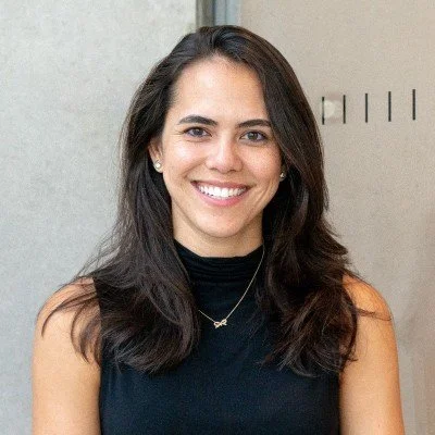 A young woman with long dark hair smiling, wearing a black sleeveless top and a delicate necklace, standing in front of a light-colored wall.