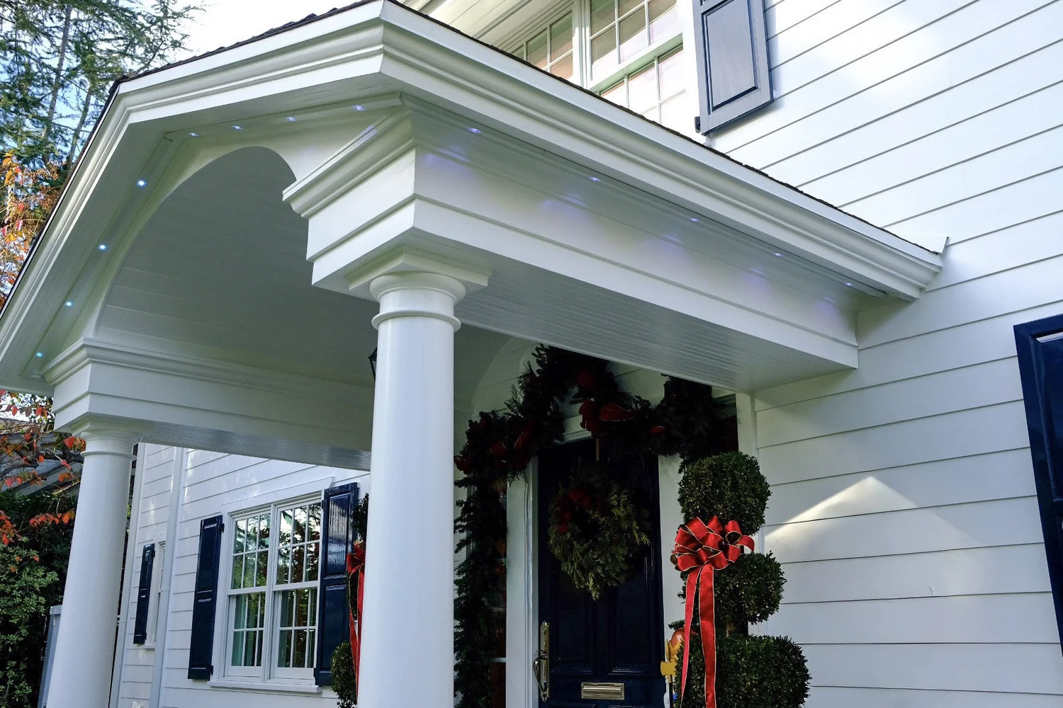 Permanent LED downlighting installed along a covered entry porch ceiling on a white colonial home