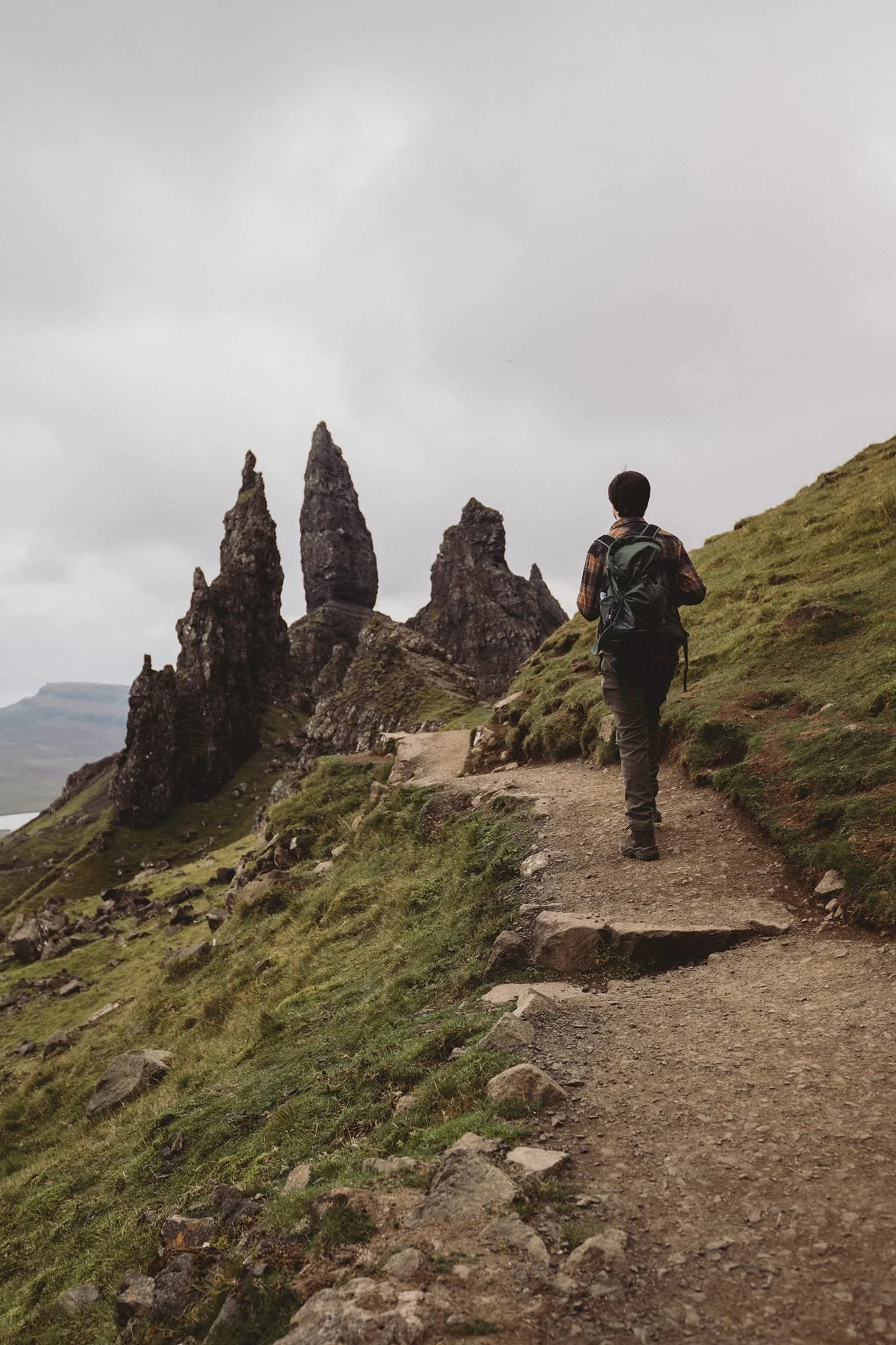 Hiker walking along a trail towards rock formations on a hillside, with overcast sky