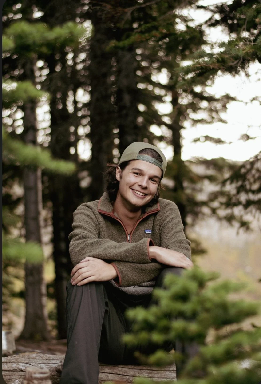 Cayden Cooke, owner of Highline Horizons, smiling outdoors in a forest, wearing a green jacket and a backwards baseball cap, sitting on a log surrounded by trees