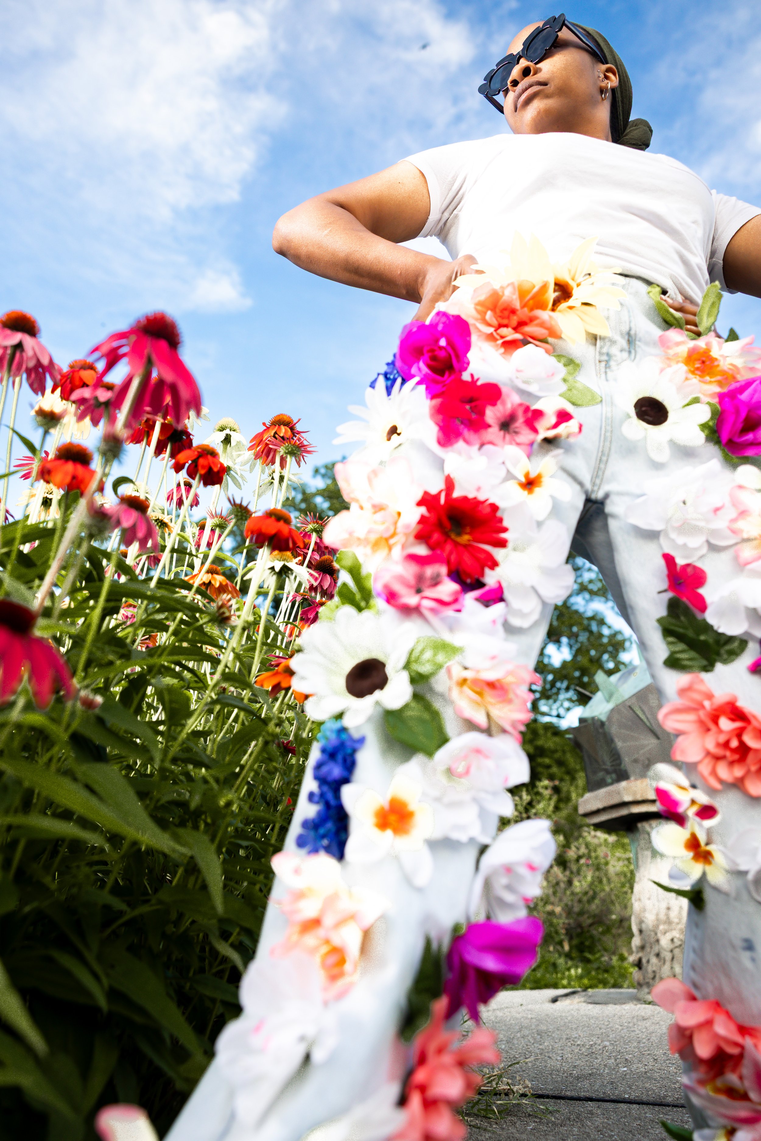 Woman standing in a flower garden with a creative outfit made of various colorful artificial flowers, viewed from a low angle against a blue sky.
