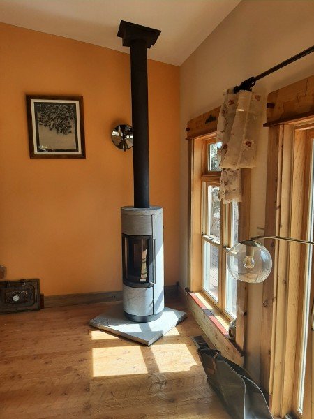 A modern cylindrical stove with a black chimney pipe connected to the ceiling in a cozy living room with wooden floors and big windows with curtains.