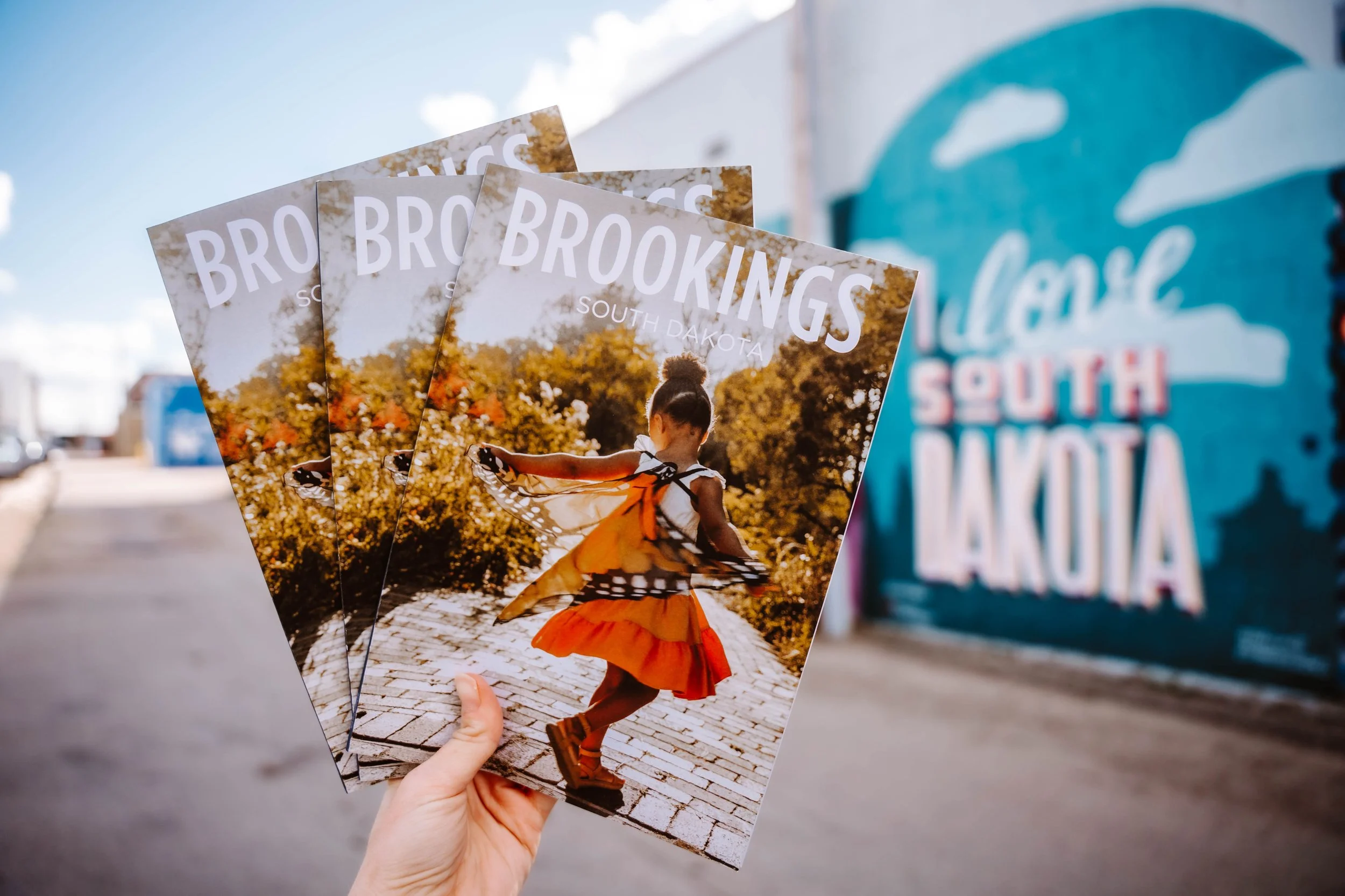 Person holding three brochures for Brookings, South Dakota, with a mural in the background.