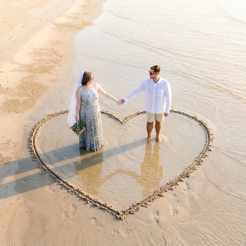 A couple standing inside a heart shape drawn in the sand at the beach, holding hands and facing each other during sunset or sunrise.