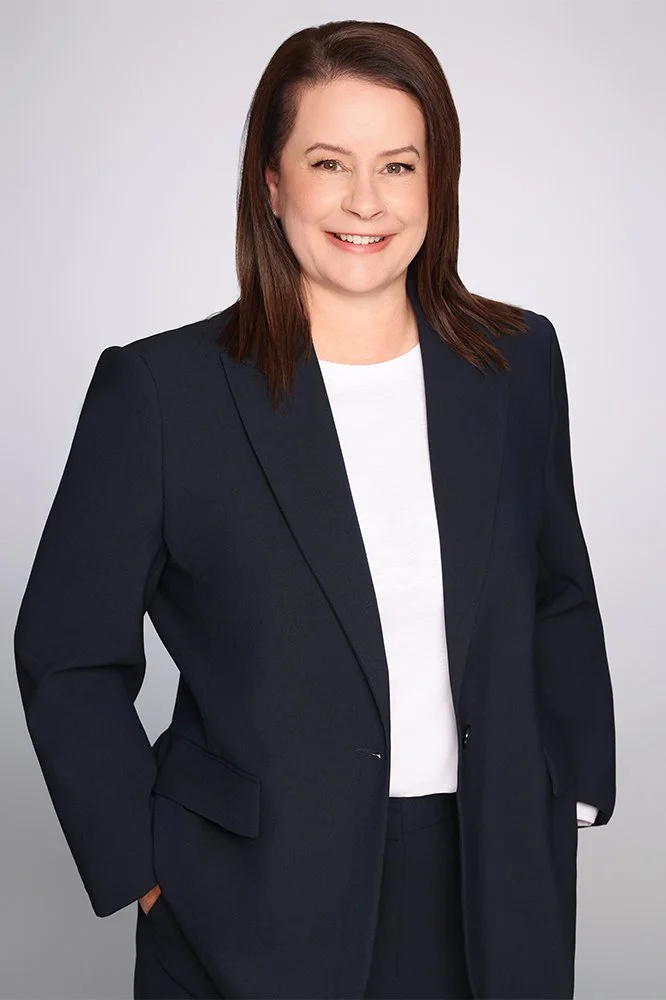 A woman with shoulder-length brown hair and fair skin, wearing a black blazer over a white shirt, smiling against a plain gray background.