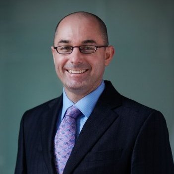 Portrait of a smiling man in a suit and tie wearing glasses against a green background.
