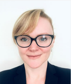 Close-up portrait of a woman with short blonde hair, wearing glasses and a black blazer, smiling against a plain white background.
