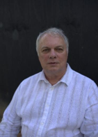 A man with short, gray hair wearing a white, striped button-up shirt standing outdoors against a dark background.