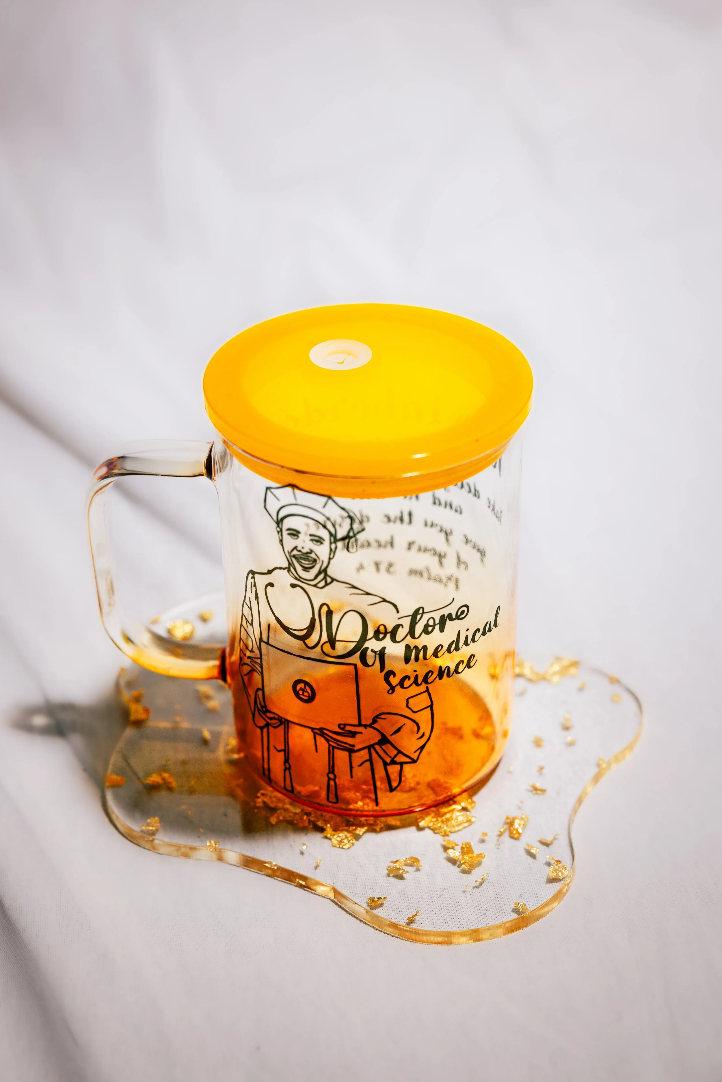 A clear glass mug with a yellow lid, featuring a doctor illustration and text that reads 'Doctor of Medical Science.' The mug is placed on a white surface with gold confetti and a gold-colored wavy ribbon around it.