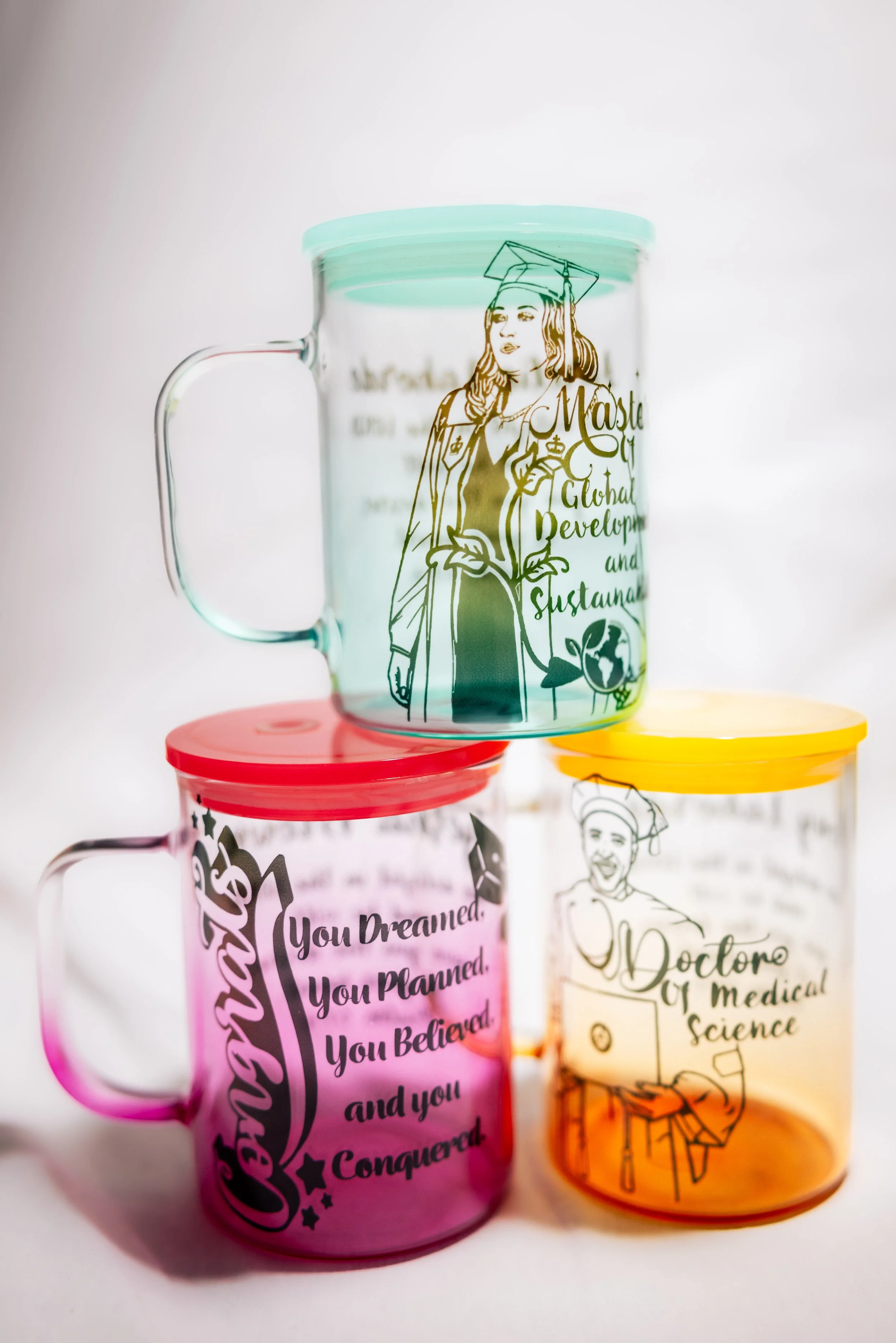 Three colorful glass mugs with inspirational and educational messages, stacked on a white surface. The top mug is transparent with green accents and features a line drawing of a graduate woman and the text "Master of Global Development and Sustainabl