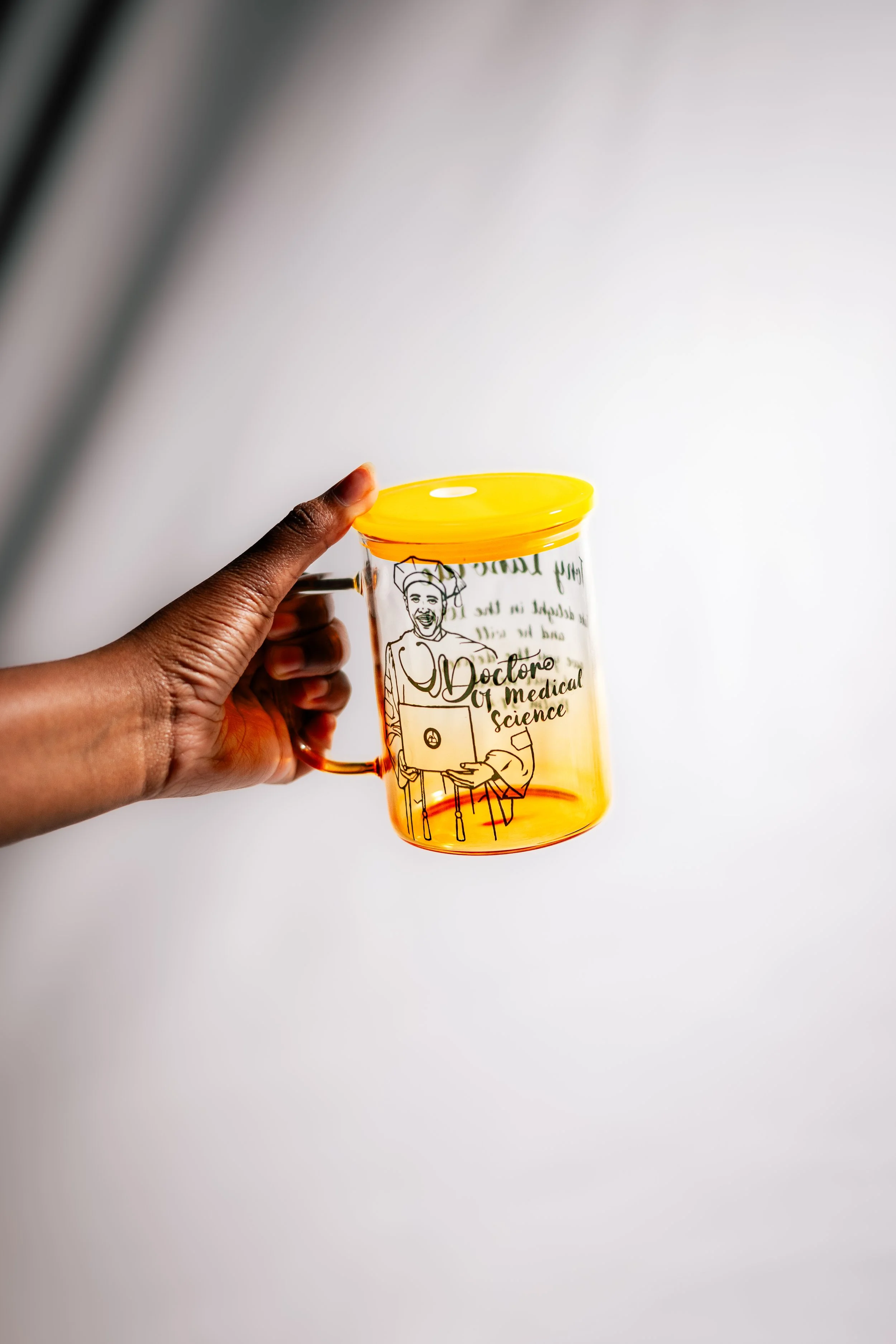 A person's hand holding a transparent orange plastic mug with a yellow lid. The mug has a cartoon illustration of a doctor and the words 'Doctor of Medical Science' printed on it.
