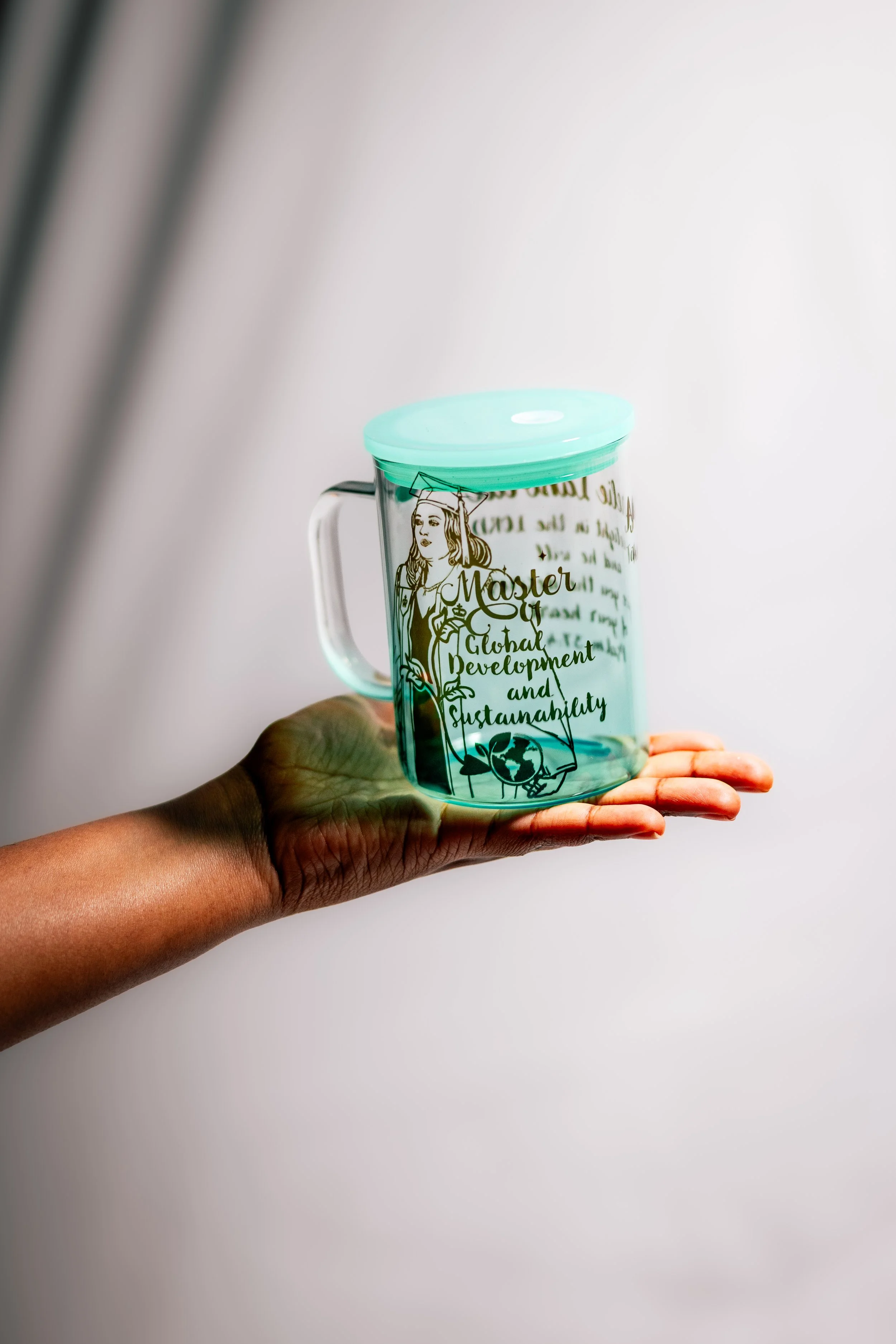 Hand holding a clear glass mug with a teal lid, decorated with the text 'Master of Global Development and Sustainability' and an illustration of a woman in a graduation cap.