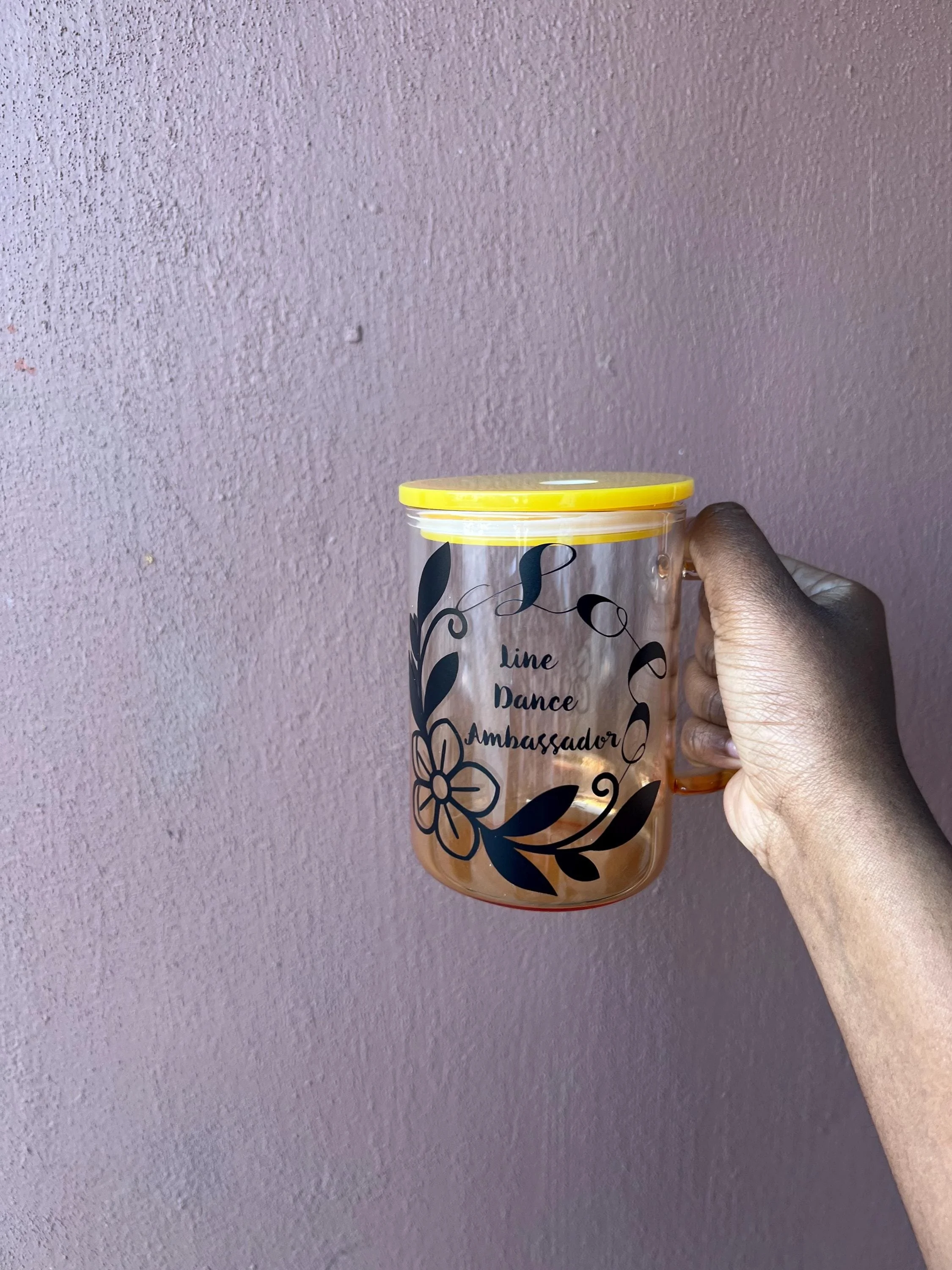 Hand holding a transparent plastic tumbler with a yellow lid, decorated with black floral design and the words "Line Dance Ambassador" written on it, against a pink textured wall background.