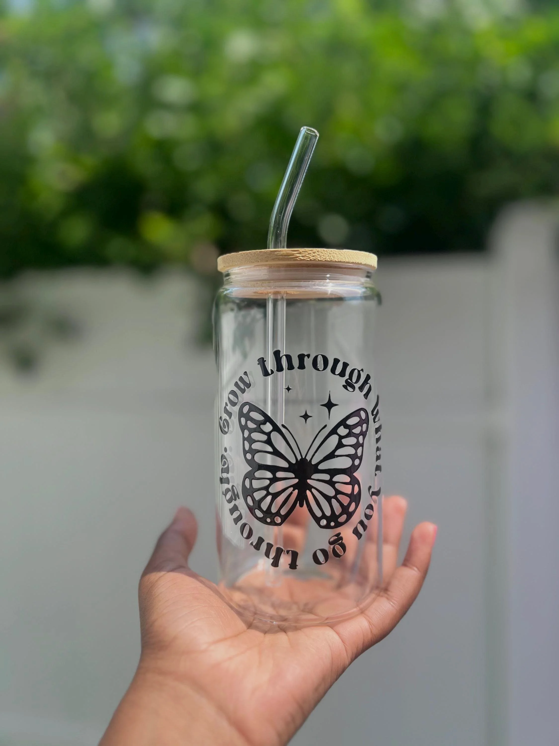 A clear glass drink container with a butterfly design and the words "log out through" on it, held by a person's hand against a blurred green background.