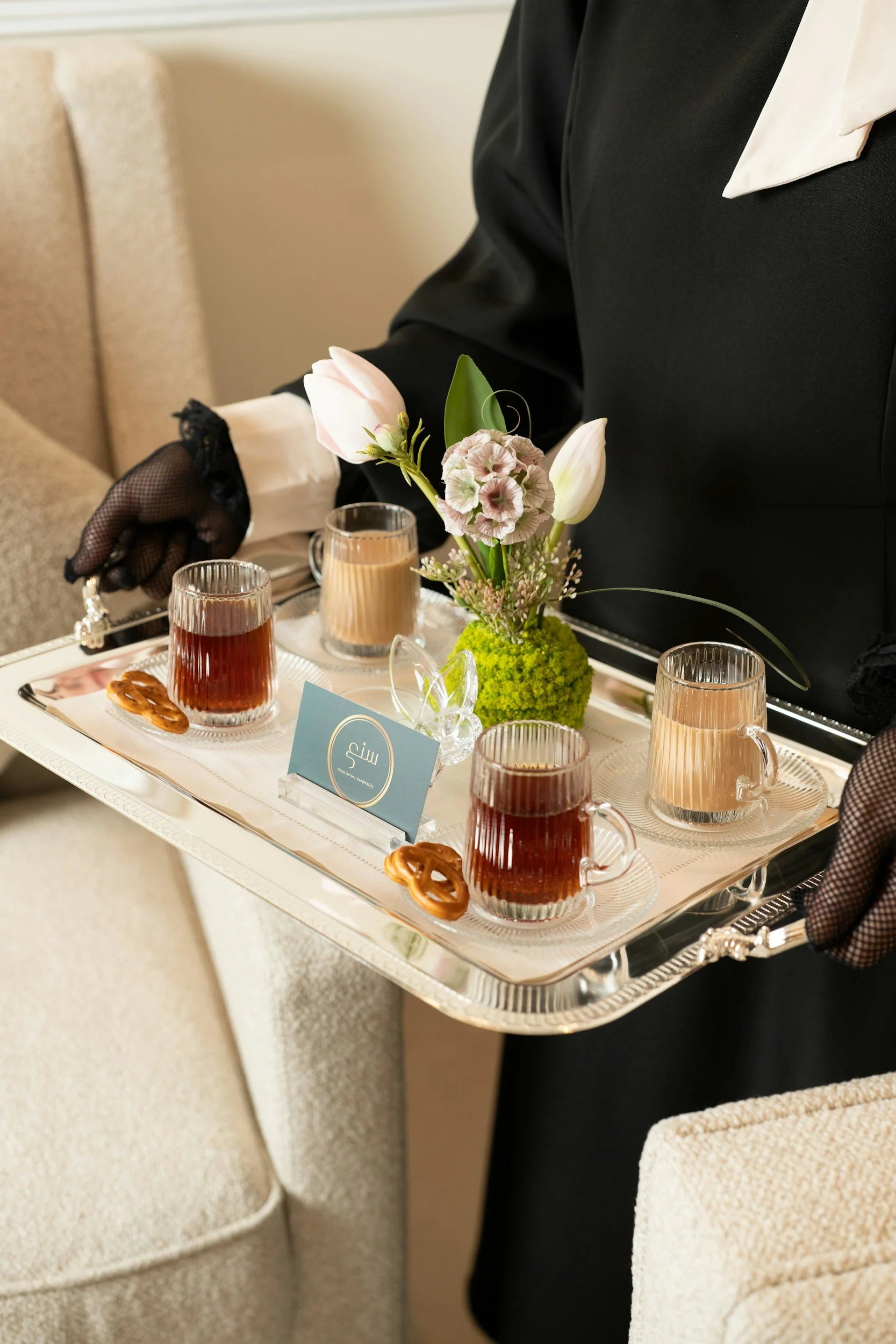 A person dressed in formal black attire with a white collar holds a silver tray with a floral arrangement of pink and white flowers, three glasses of tea, and small pretzels. The tray has a blue card with a white logo and text.