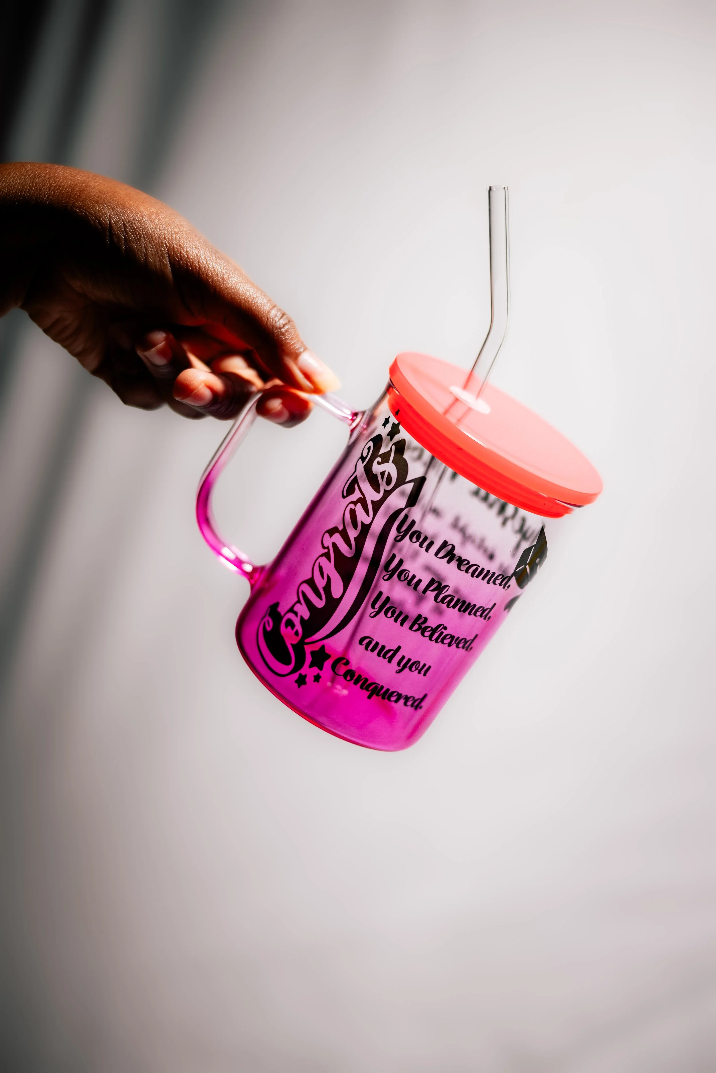 A person holds a pink tumbler with a motivational quote written on it. The tumbler has a red lid and a clear, flexible straw. The person's hand is visible, and the background is mostly white with some shadowed areas.