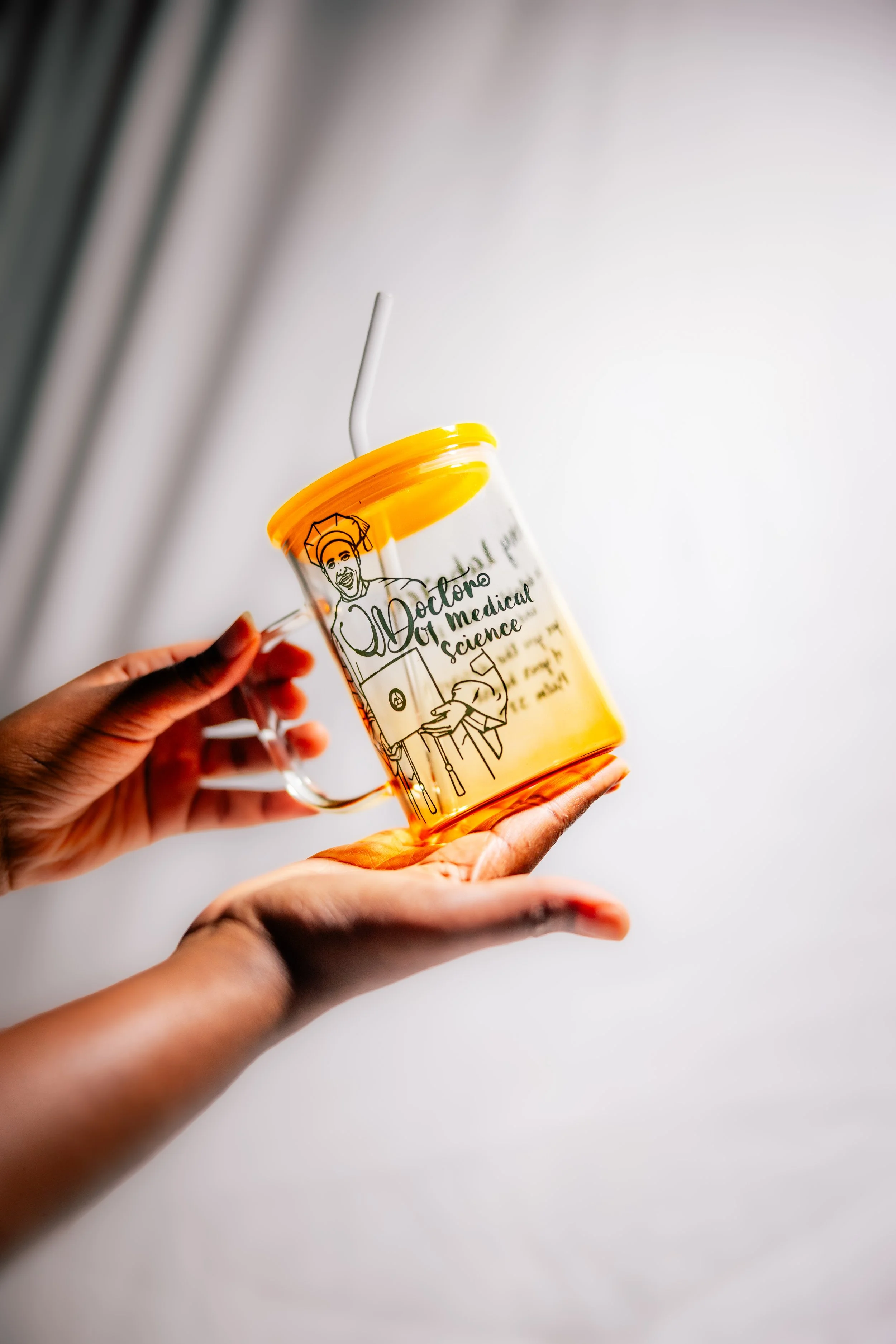 A hand holding a transparent orange cup with a white straw. The cup has a black illustration of a doctor and the words 'Doctor of Medical Science' written on it.