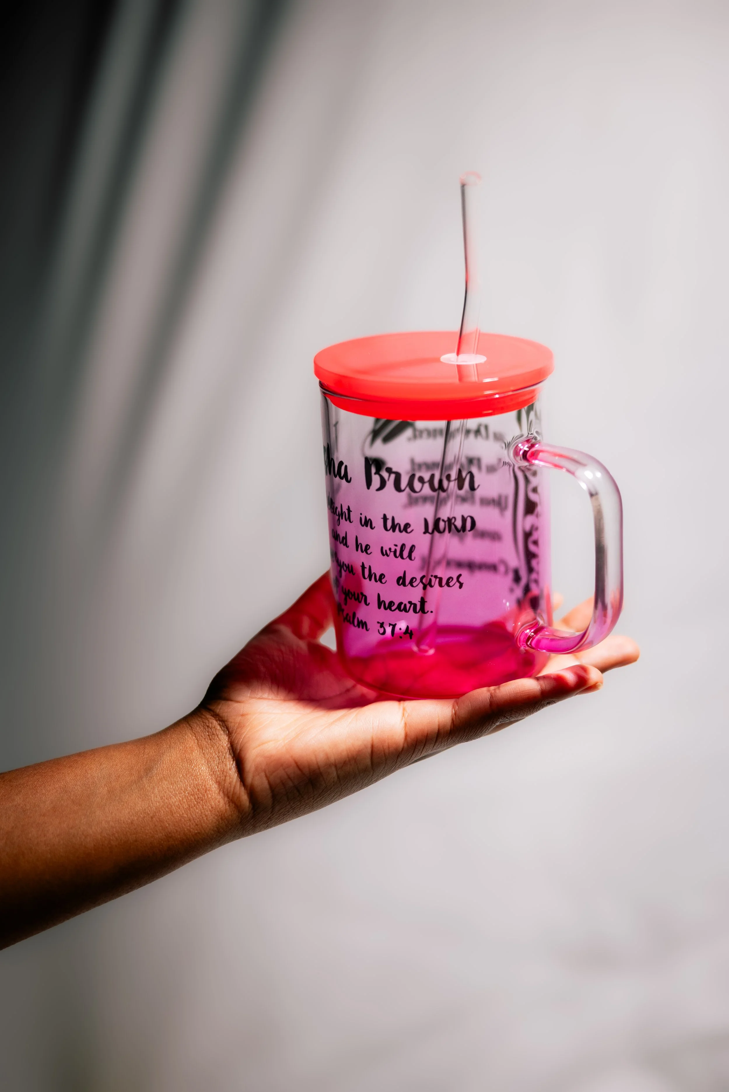 A person holding a pink transparent mug with black text on it, with an upward moving red and black straw inside the mug. The person's hand and part of their arm are visible. The background is plain and light-colored.