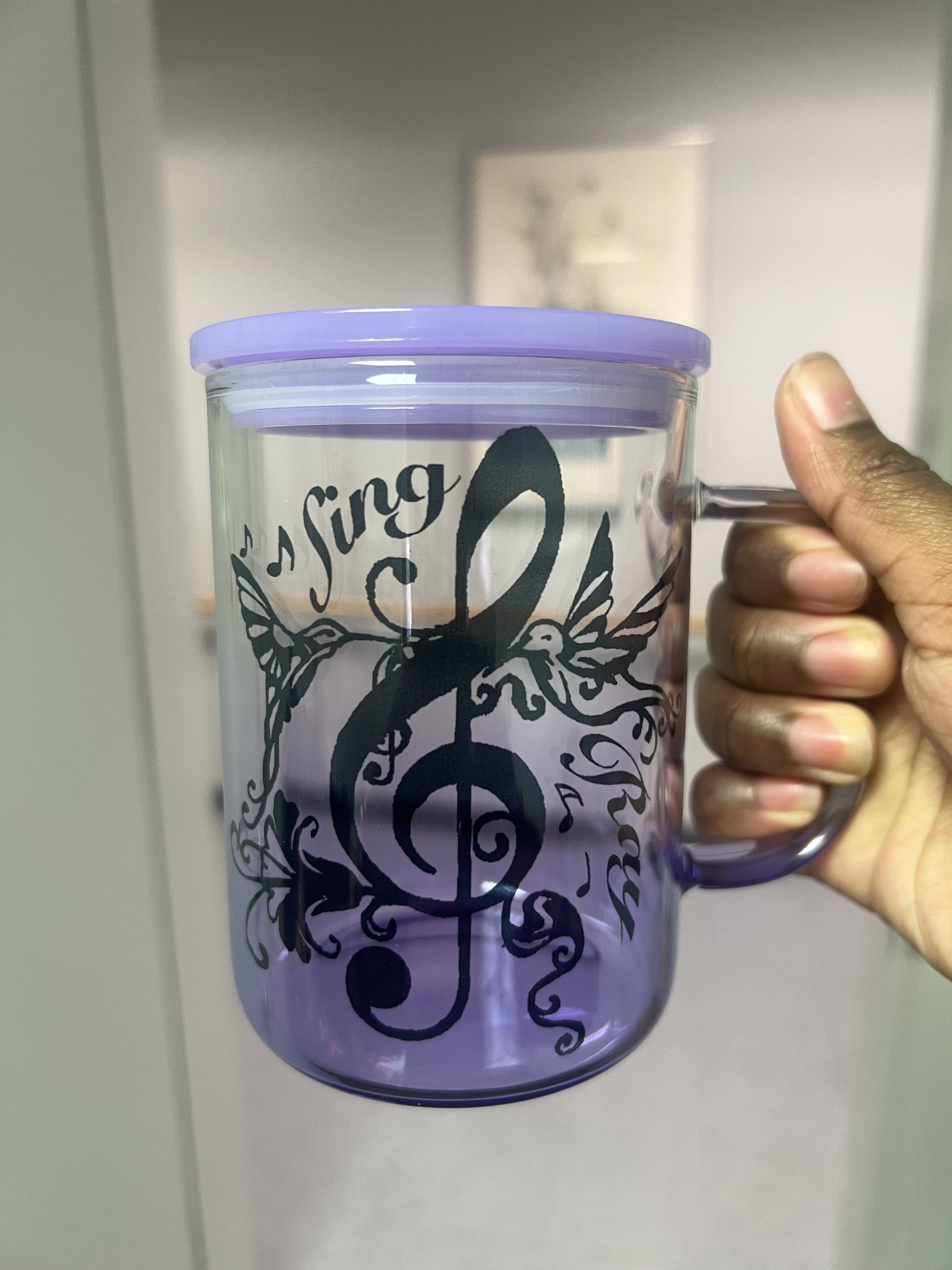 A clear plastic tumbler with a purple lid being held by a person's hand, featuring black text and design, possibly with a floral or ornate motif.