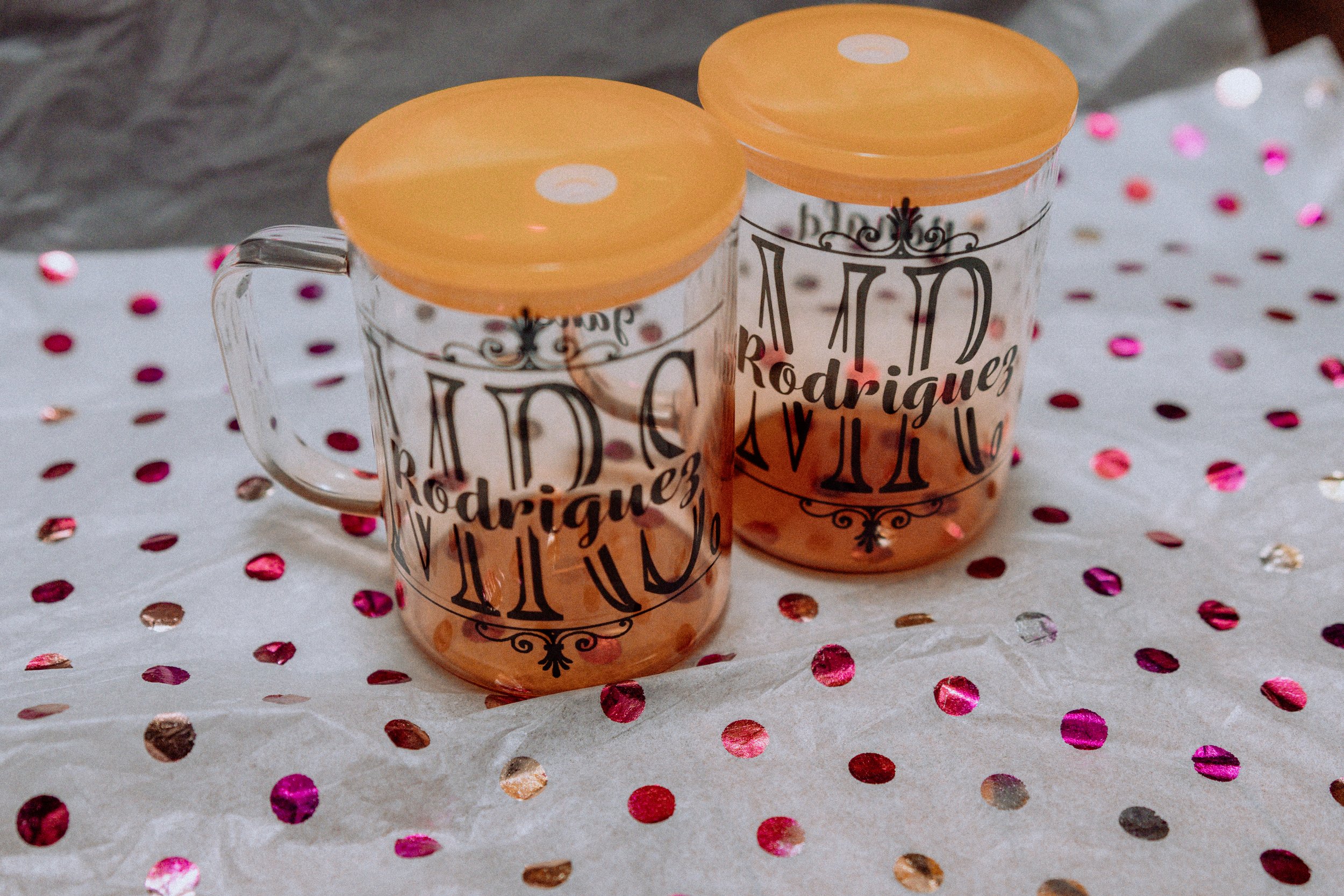 Two glass mugs with orange lids and black text, placed on a white surface decorated with pink, red, and silver confetti.