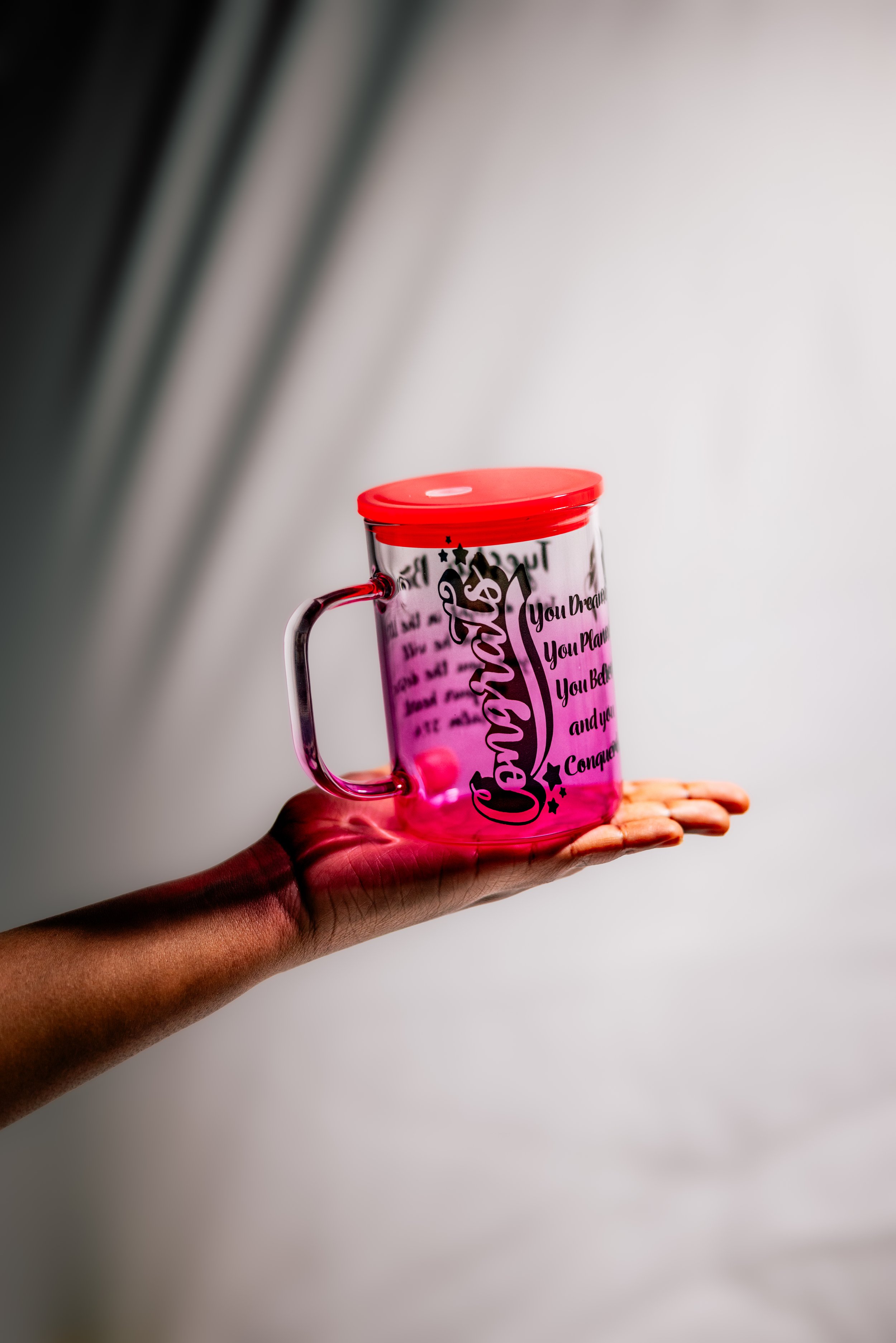 A person's hand holding a transparent pink mug with a red lid, against a plain background.