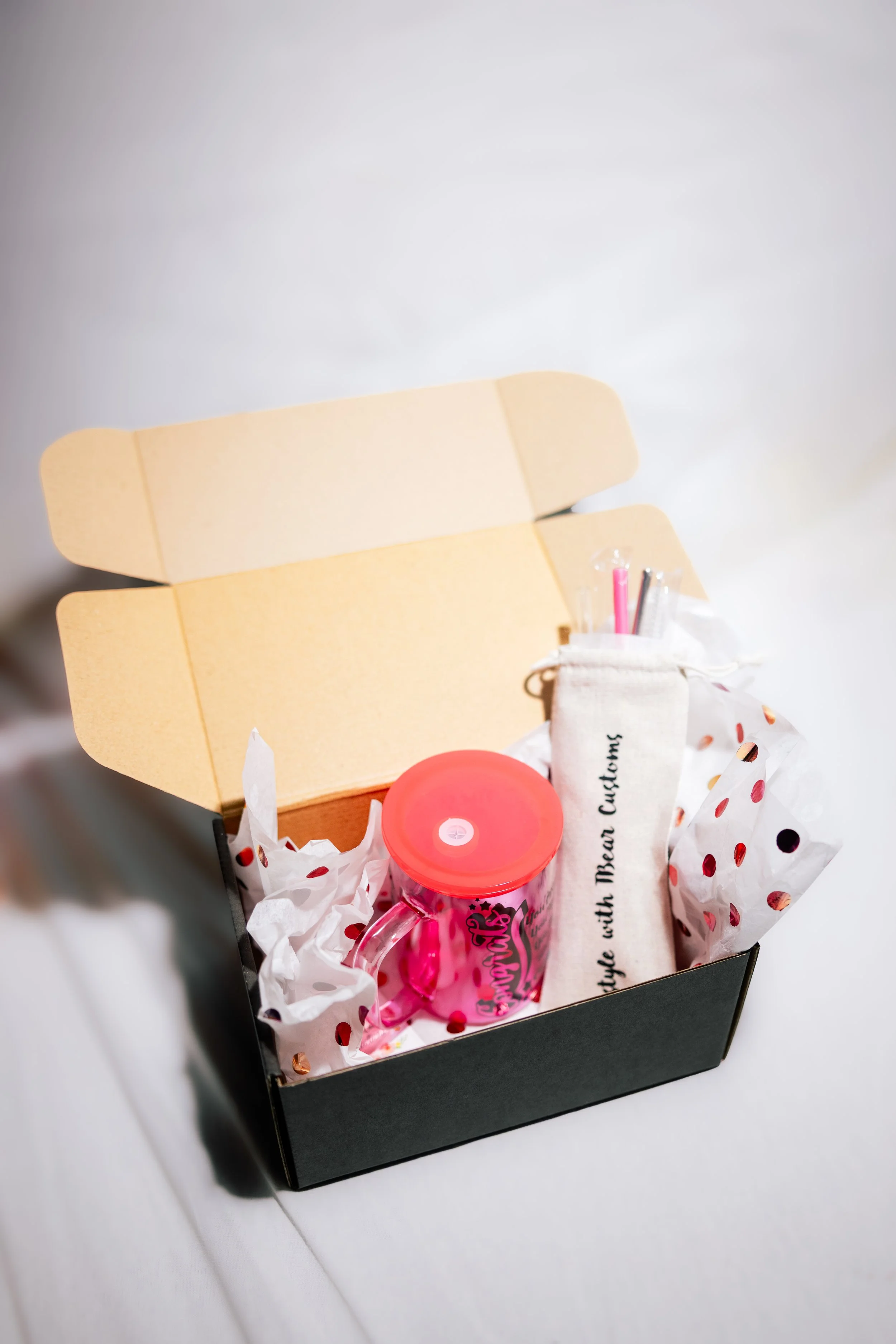 Black gift box with tissue paper, containing a pink drink, a small pouch, and yellow envelopes for note cards, on a white surface.