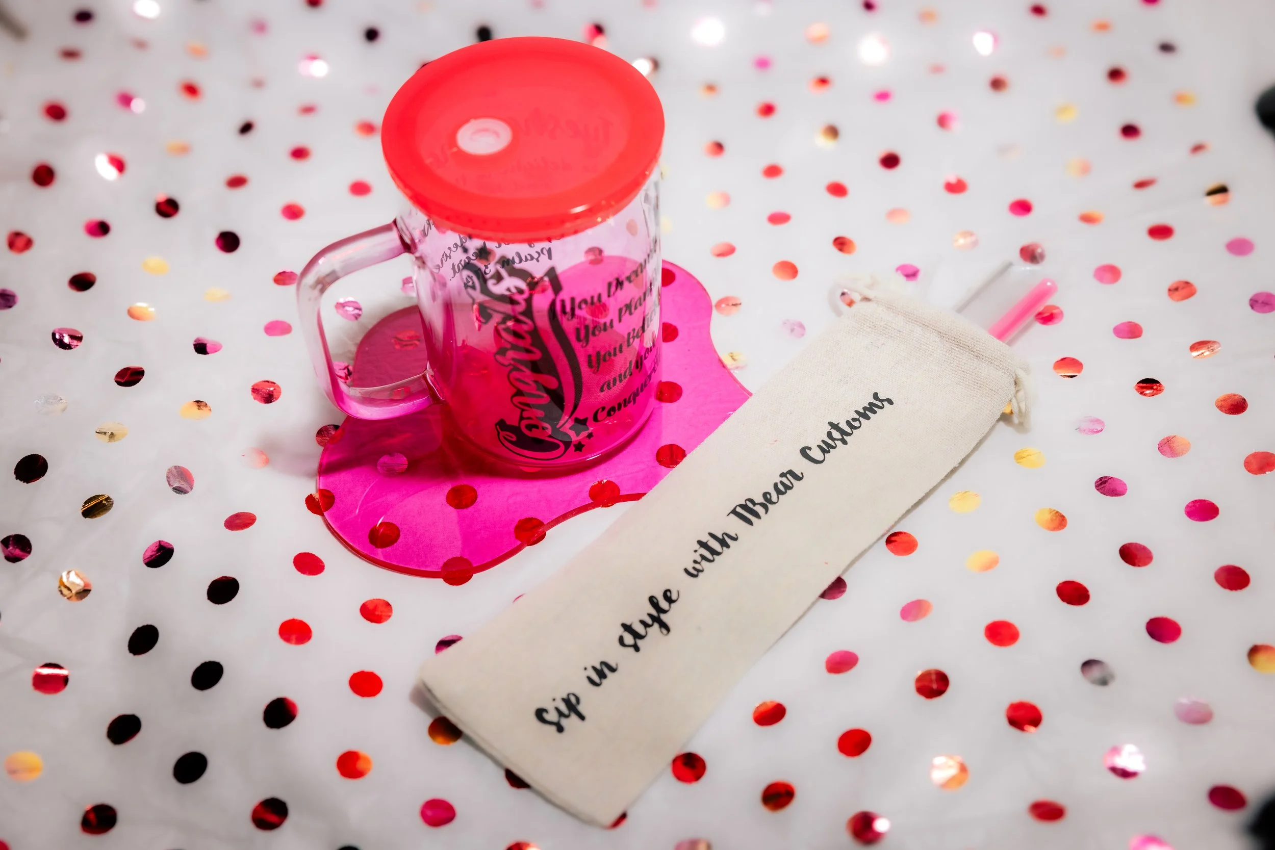Pink glass mug with black text, topped with a red lid, placed on a pink paper heart, on a metallic gold and pink polka-dot background, accompanied by a white cloth and pink straw